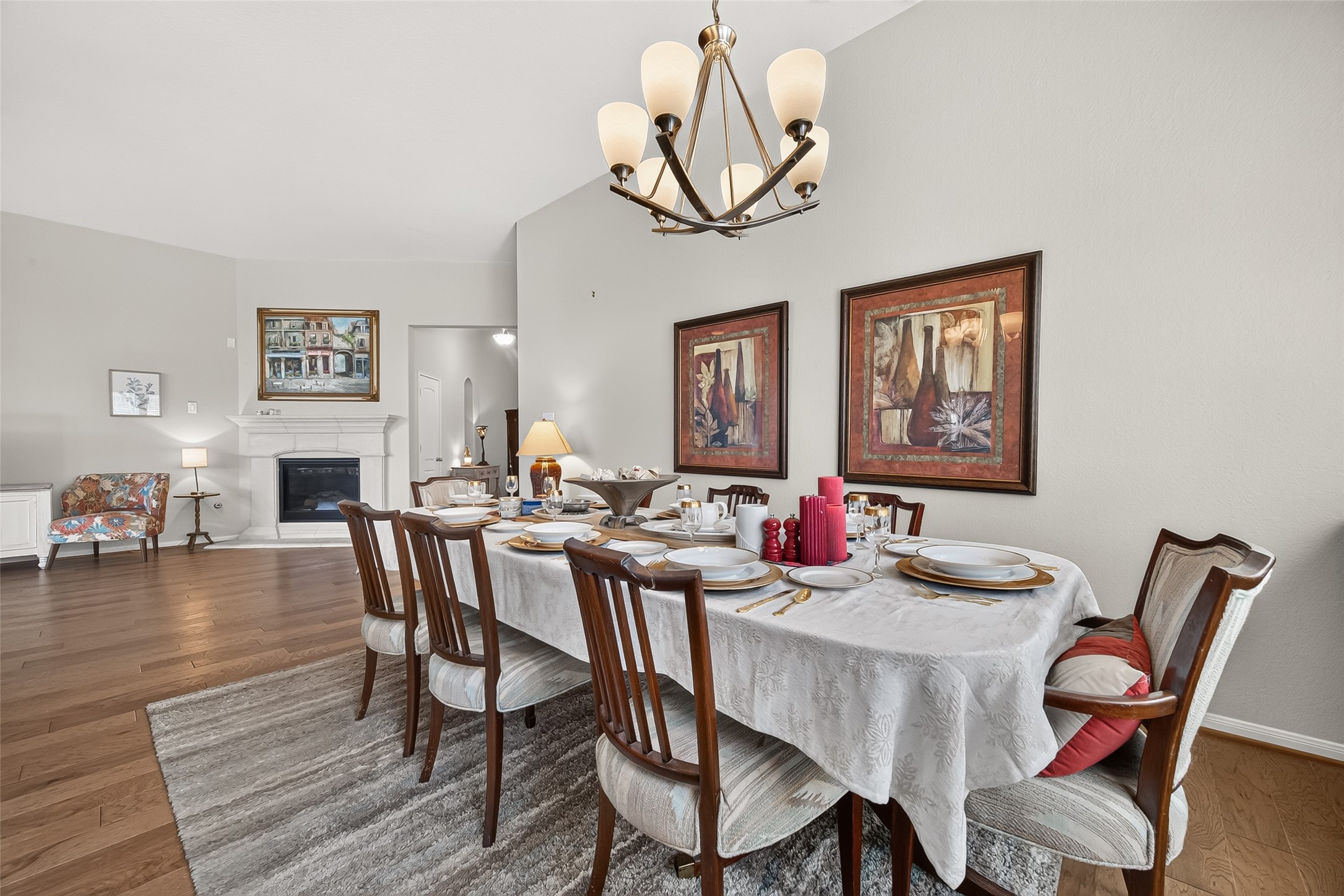 4910 Culver Lane Rosenberg, TX 77471 - Photo 7 of 50 The formal dining room features a large, elegant table set for eight, with a stylish chandelier overhead. The space is complemented by warm wood flooring, a cozy fireplace, and tasteful artwork, creating an inviting atmosphere for family gatherings.