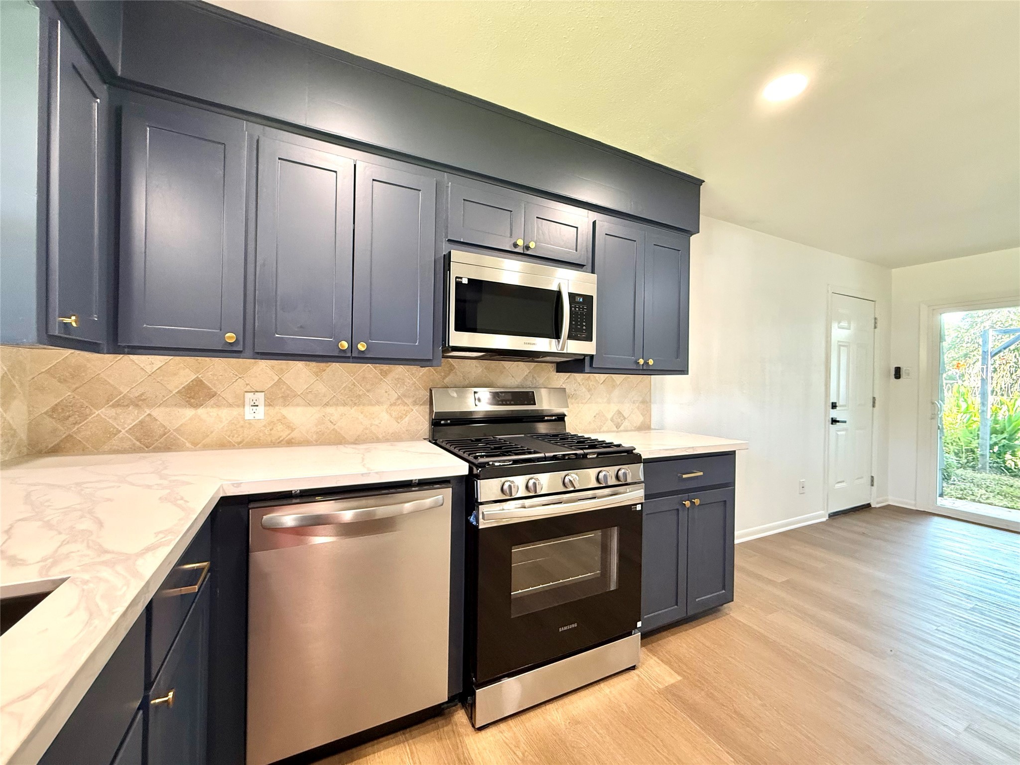 129 Chestnut Street Lake Jackson, TX 77566 - Photo 14 of 20 This kitchen features elegant blue cabinetry with gold hardware, a modern stainless steel stove and microwave, and a sleek dishwasher. The space is complemented by marble-look countertops and a stylish tiled backsplash, offering a bright and inviting atmosphere.