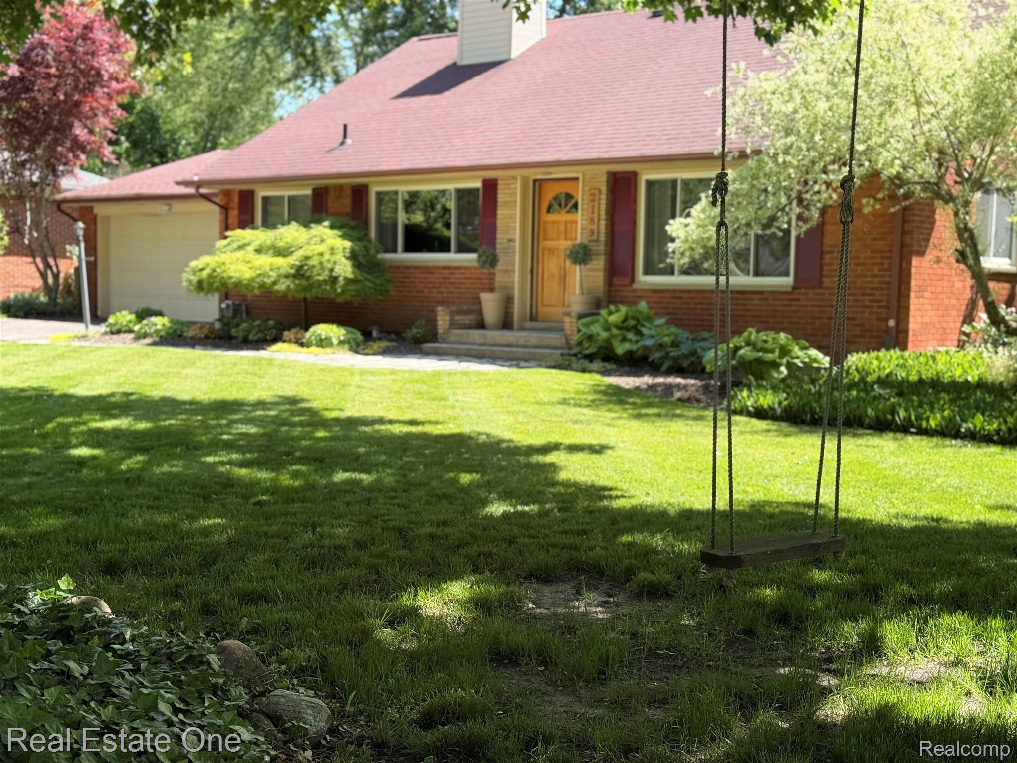 Brick facade residence featuring a red shingle roof, attached single-car garage, and a lush front lawn with mature trees