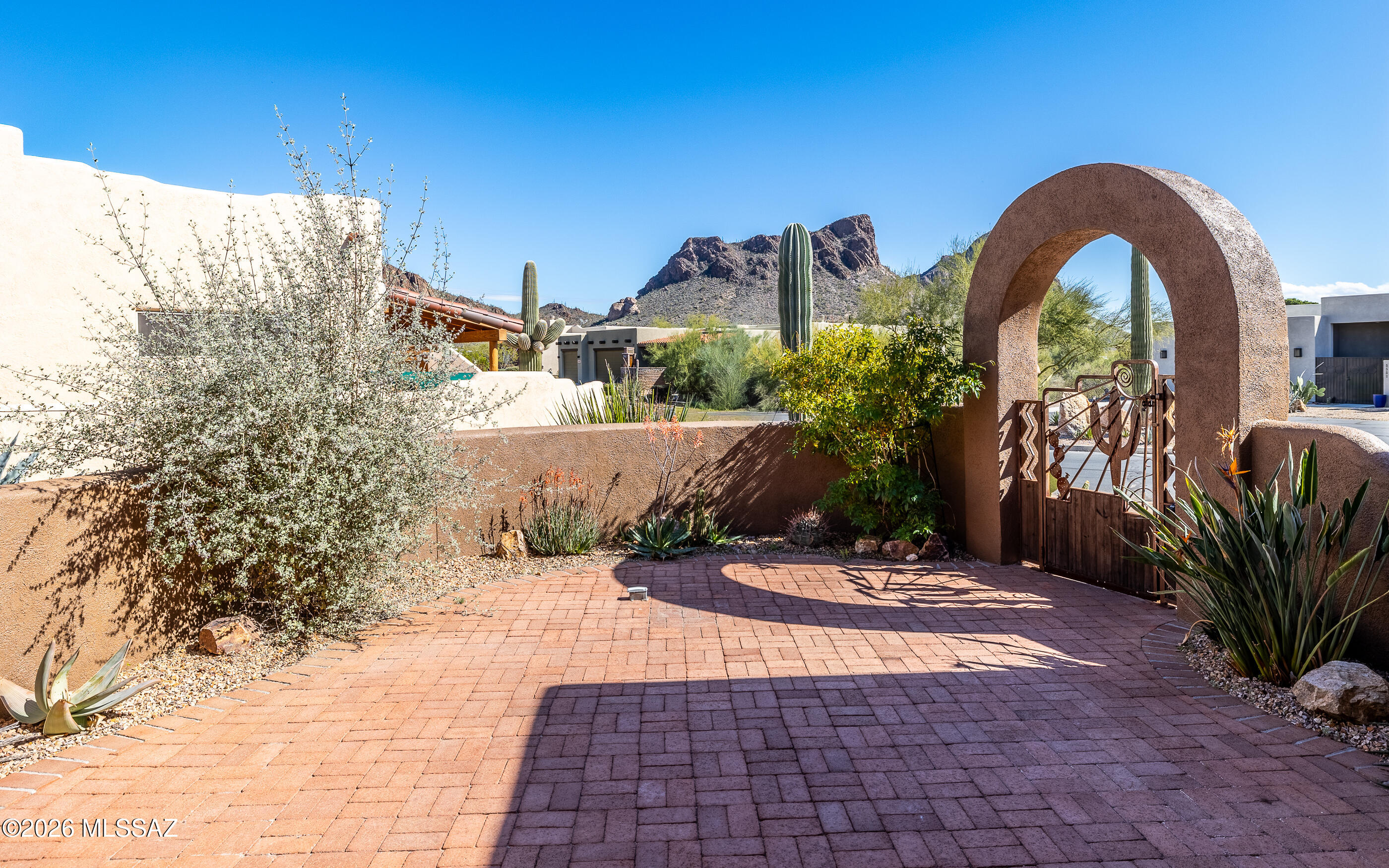 5550 West Open R Place Tucson, AZ 85713 - Photo 4 of 48 Gorgeous reddish-brown pavers, create a warm, textured walkway that leads through an arched adobe-style gateway, with views of the rugged mountain peaks rising under a clear blue sky!
