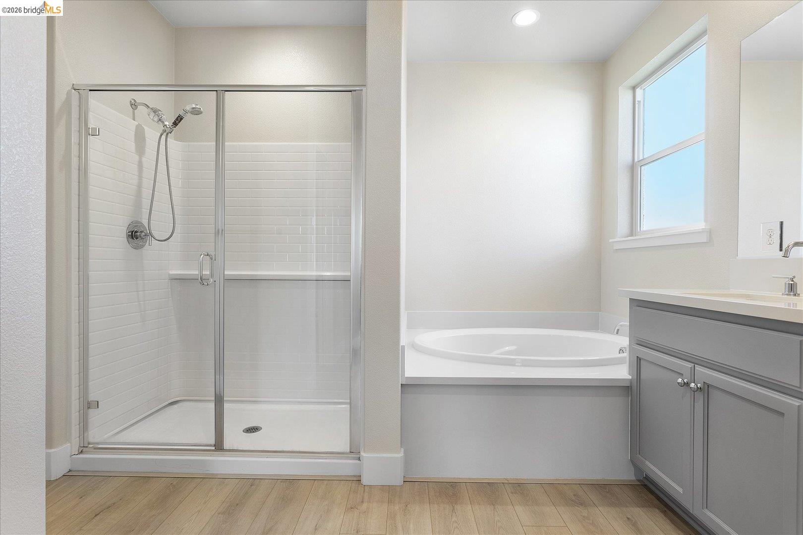 5601 Ridgepoint Way Antioch, CA 94531 - Photo 40 of 59 Contemporary bathroom featuring a glass-enclosed shower with subway tile, a built-in soaking tub, a double vanity with gray cabinetry, a window, and wood-finish flooring