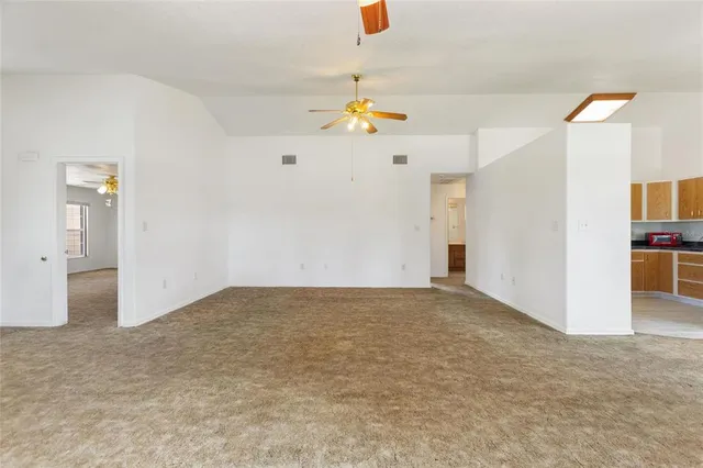 an empty room with chandelier fan and kitchen view