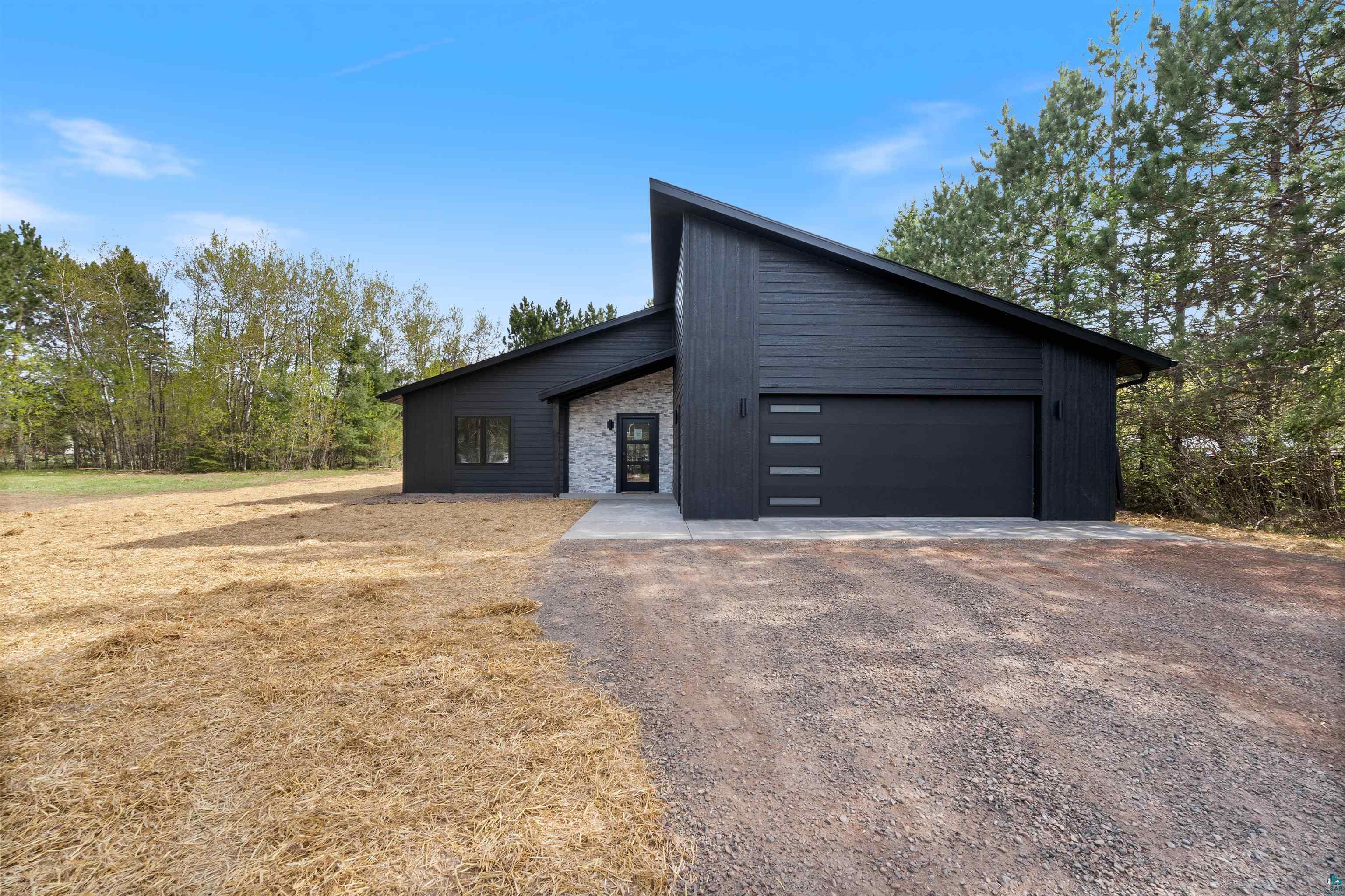 1386 Stephen Road Cloquet, MN 55720 - Photo 1 of 25 Modern exterior featuring dark vertical and horizontal siding, a striking sloped roofline, a gravel driveway, a contemporary garage door with horizontal windows, and a stone facade entry