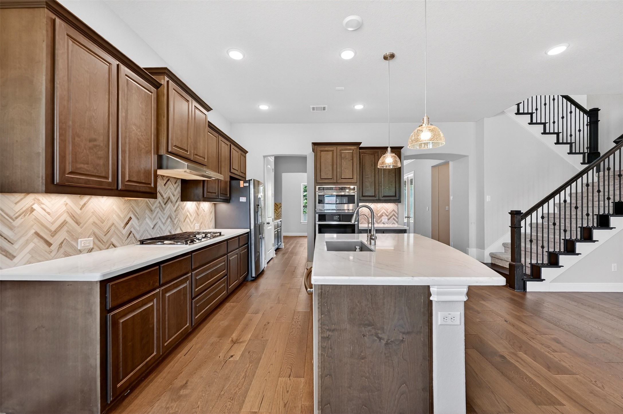 1201 Positano Pass Leander, TX 78641 - Photo 12 of 40 Spacious kitchen featuring an extended island with a built-in sink, wood-finish flooring throughout, stainless steel appliances, and dark wood cabinetry with a herringbone pattern tile backsplash