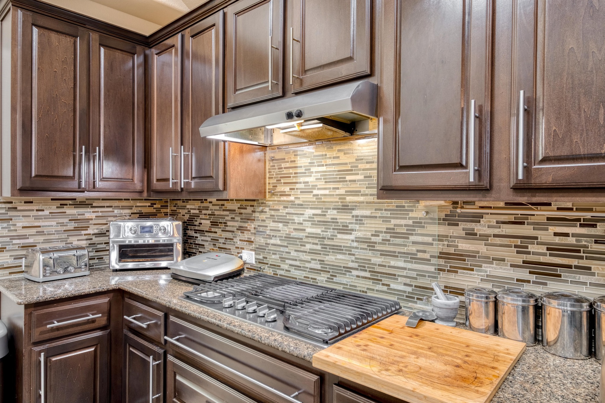 3040 Sachen Street Georgetown, TX 78626 - Photo 11 of 40 Kitchen featuring under cabinet range hood, backsplash, stainless steel gas cooktop, dark brown cabinets, and light stone countertops