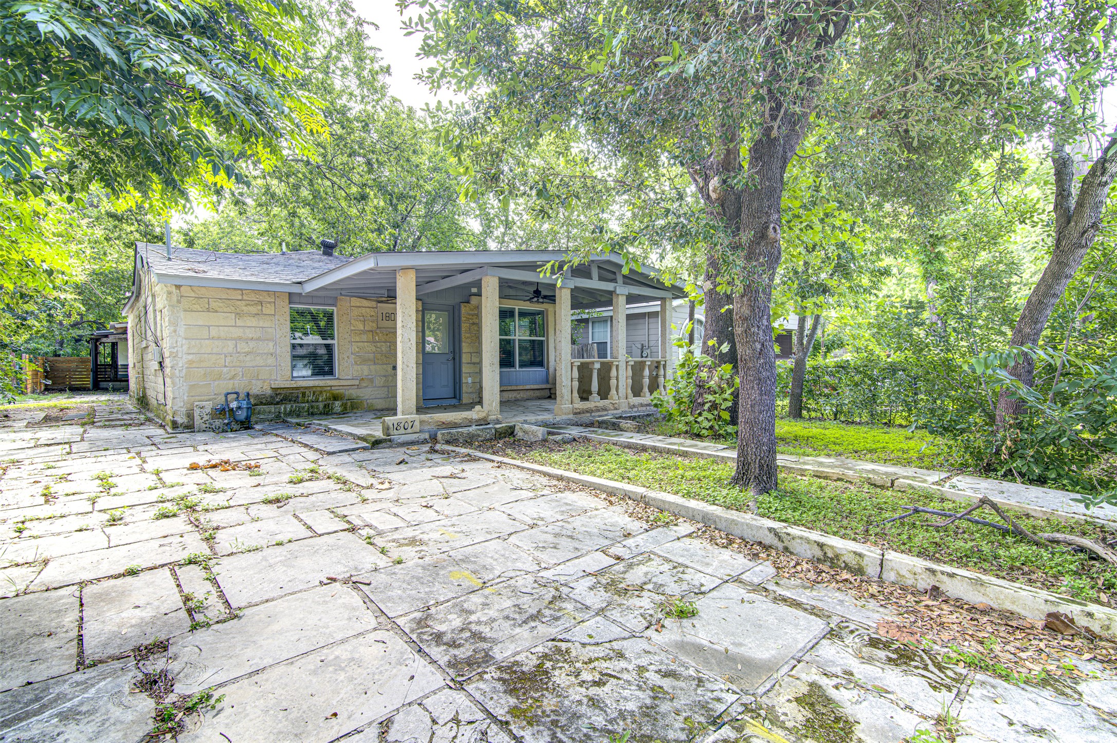 Mature trees frame a cozy stone home, featuring a welcoming covered porch and an expansive paved driveway.