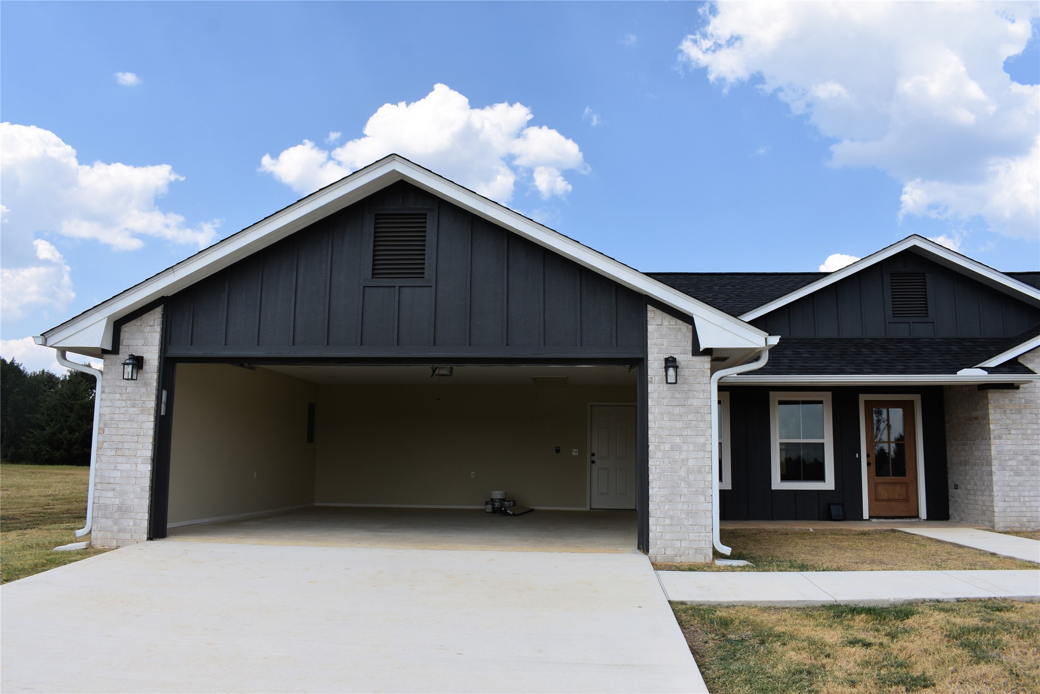 2915 East 2090 Road, Unit LOT 1 Hugo, OK 74743 - Photo 24 of 28 Contemporary exterior featuring white brick accents, dark vertical siding, a wood-finish front door, a two-car carport, and a sidewalk