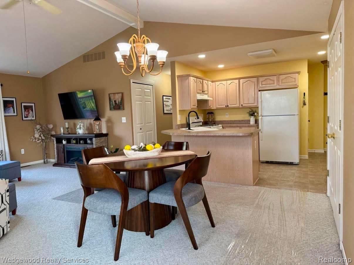 Vaulted ceiling with exposed beams, recessed lighting, wood-finish kitchen cabinetry, breakfast bar, and an electric fireplace with stone-finish surround