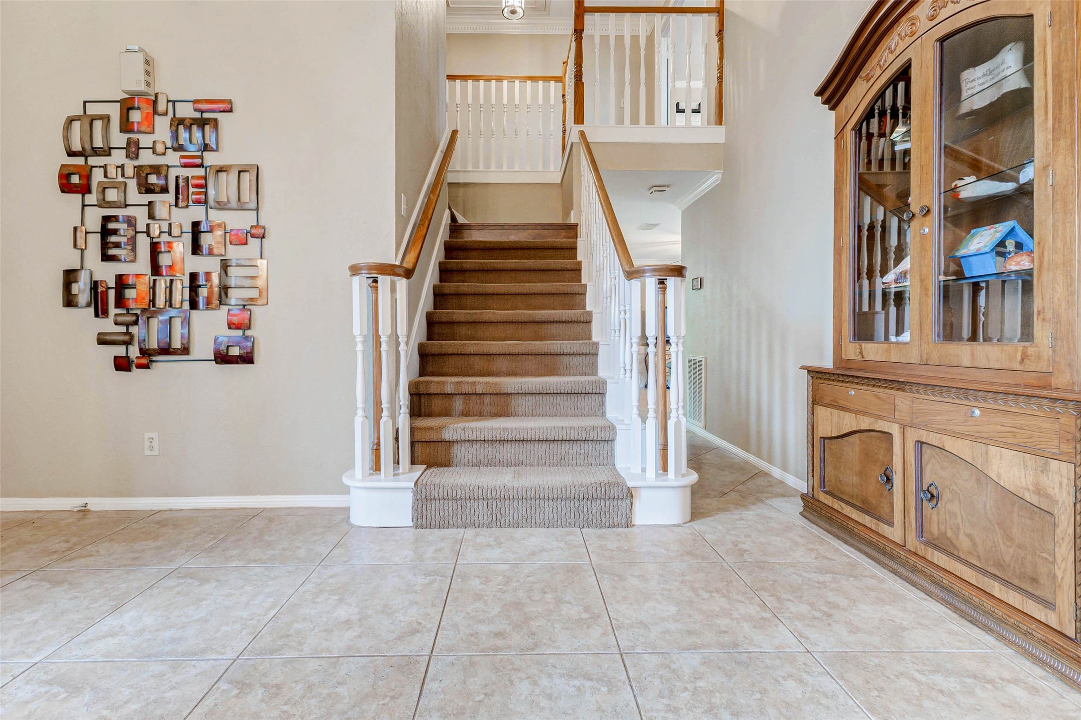 15811 Spring Forest Drive Houston, TX 77059 - Photo 13 of 27 This inviting foyer features a carpeted staircase with white railings leading to the upper floor. The space is enhanced by a decorative wall piece and a wooden cabinet, all set on elegant tiled flooring.