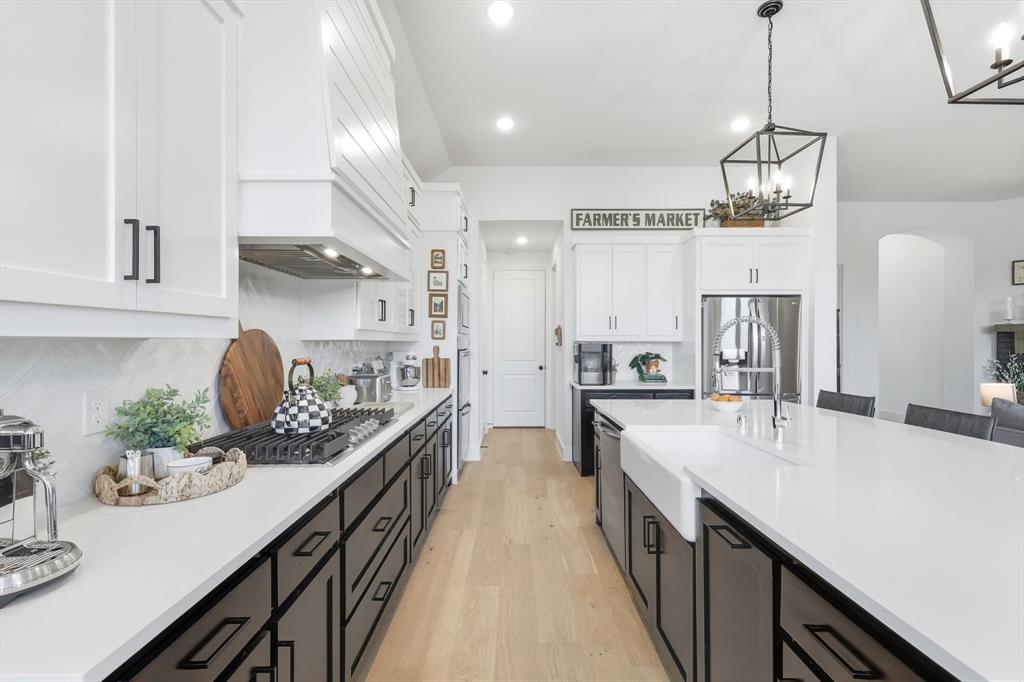 4608 Mount Vista Road Oak Point, TX 75068 - Photo 19 of 39 Contemporary kitchen featuring white shaker upper cabinetry and dark lower cabinetry with matte black hardware, light-toned wood-finish flooring, a white apron-front sink, and a large island with white countertops