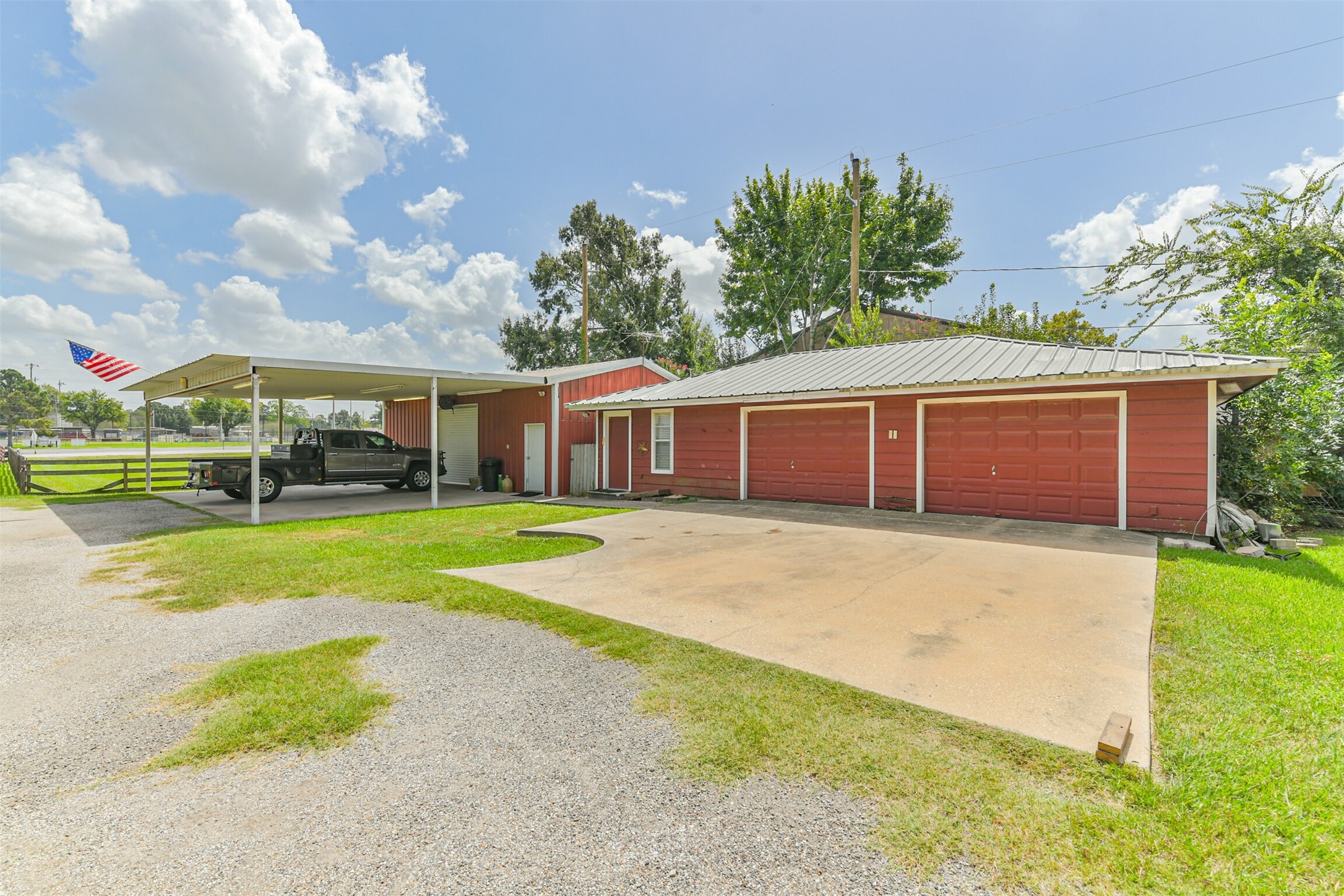 307 West Hufsmith Road Tomball, TX 77375 - Photo 4 of 44 Spacious property featuring a vibrant red garage with three doors, a covered carport, and ample driveway space, all set against a backdrop of lush greenery and clear blue skies.