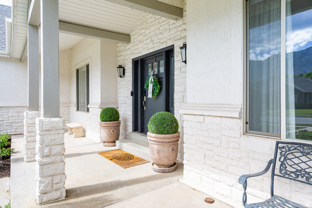 868 Braddock Lane Alpine, UT 84004 - Photo 4 of 64 Front entry featuring a dark-paneled door with opaque glass details, white stacked stone veneer, textured stucco siding, and a covered porch with stone-clad columns