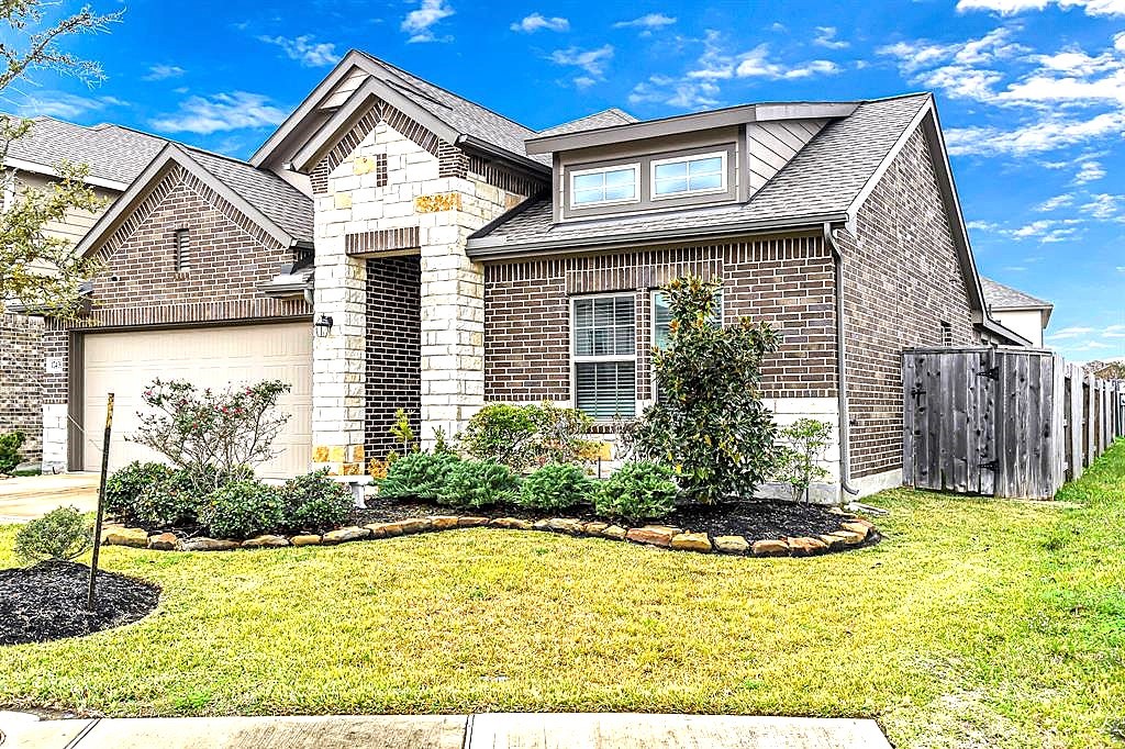 17439 Chester Valley Trail Hockley, TX 77447 - Photo 3 of 22 This charming brick home features a modern design with a gabled roof, a two-car garage, and a neatly landscaped front yard. The facade blends stone accents with traditional brickwork, set against a bright blue sky. A wooden fence lines the side, offering privacy.