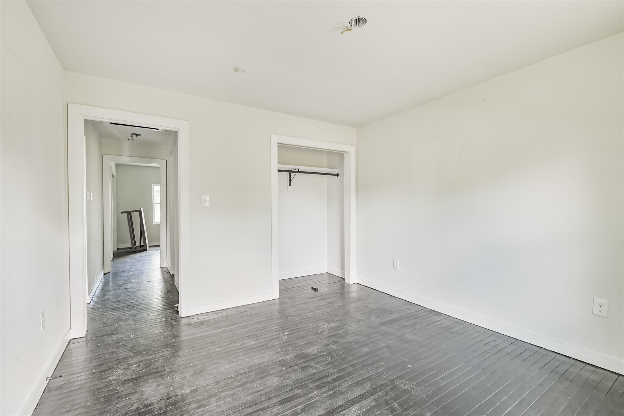 209 Leago Street Houston, TX 77022 - Photo 15 of 24 This photo shows a simple, empty room with light-colored walls, a small closet, and dark hardwood floors. There's a hallway leading to another room with natural light from a window. The space appears ready for customization or renovation.