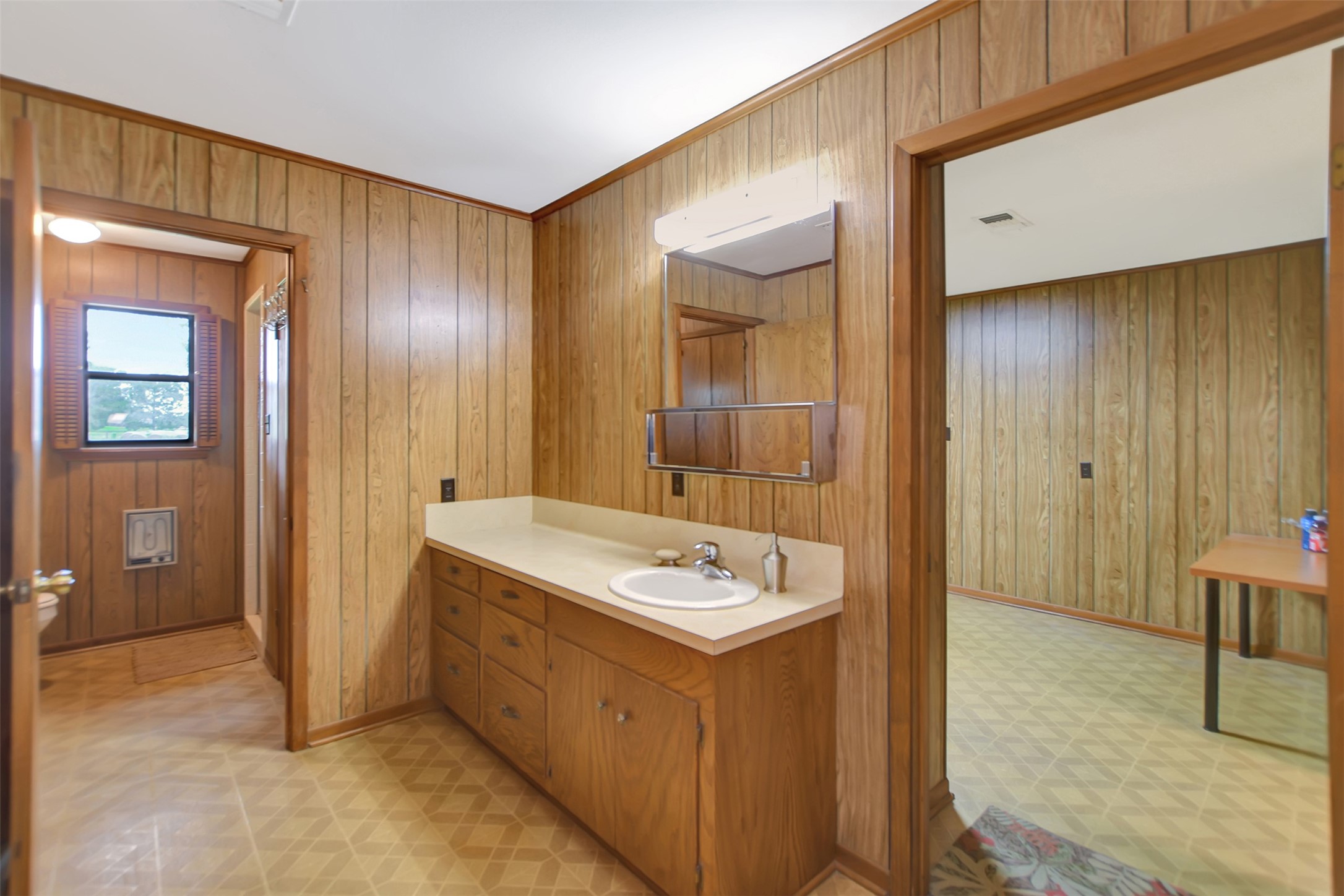 341 South S Loop Bellville, TX 77418 - Photo 18 of 33 This photo shows a retro-style bathroom area with wood paneling, a single sink vanity, and an adjacent room. The space features ample cabinetry and natural light from a window in the connected room.
