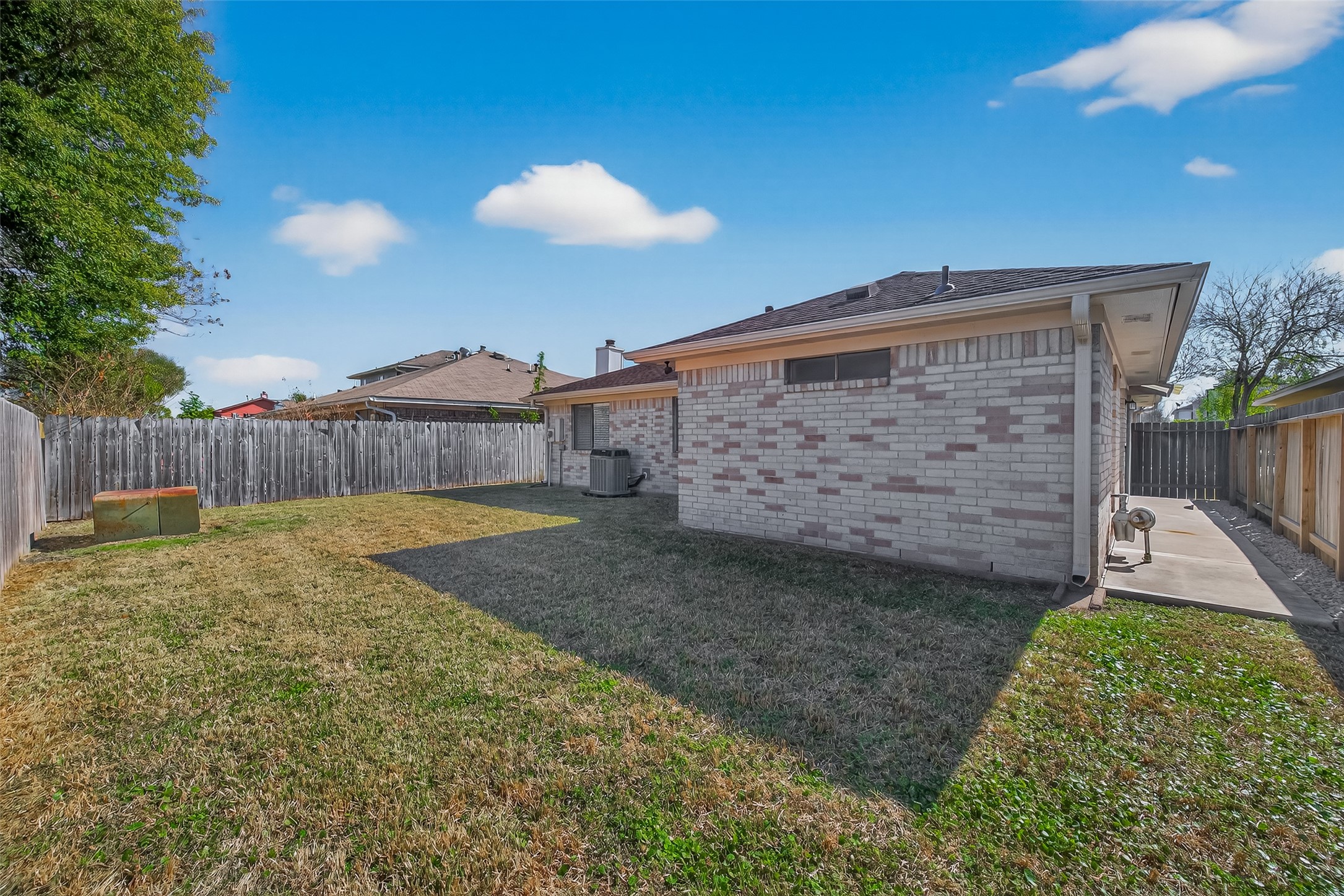 10810 Umber Court Houston, TX 77099 - Photo 42 of 43 This photo showcases a backyard with a well-maintained lawn, enclosed by a wooden fence, providing privacy. The side of a brick house is visible, with an outdoor air conditioning unit. There's a paved walkway along the house, offering easy access around the property.
