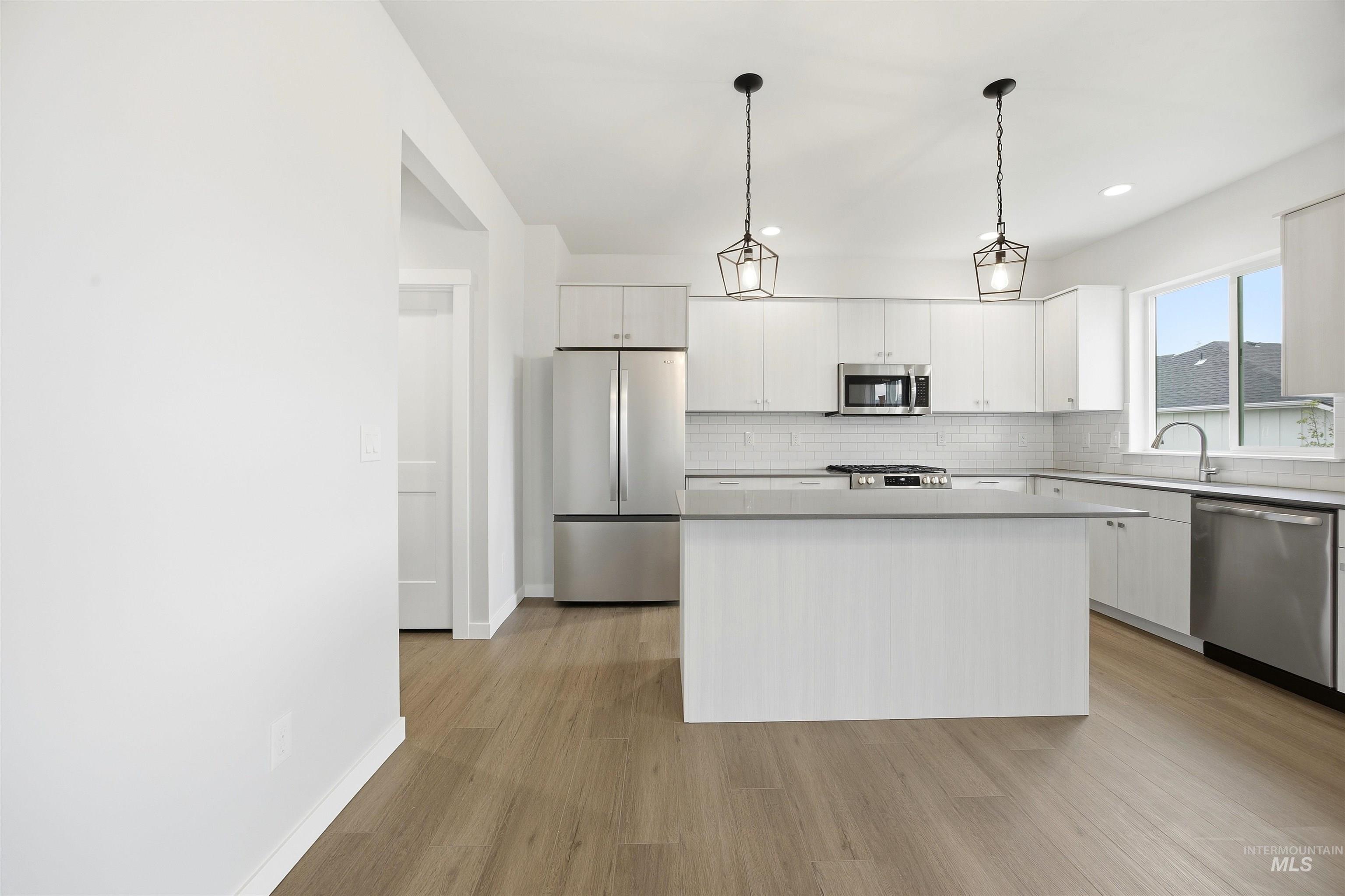 6447 South Sunfish Way Boise, ID 83709 - Photo 4 of 29 Modern kitchen featuring a central island with a light-toned countertop, white cabinetry, stainless steel appliances, wood-finish flooring, and two geometric pendant lights