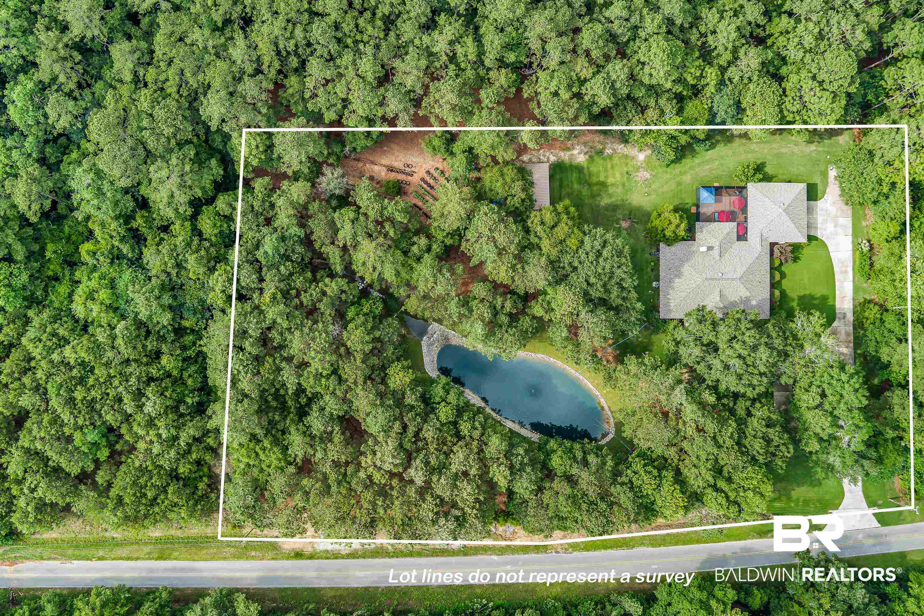 7014 Blakeley Road Daphne, AL 36527 - Photo 44 of 45 Private residential property featuring a gray-roofed home, a paved driveway, an uncovered patio area, a pond with a water feature, and a surrounding wooded landscape