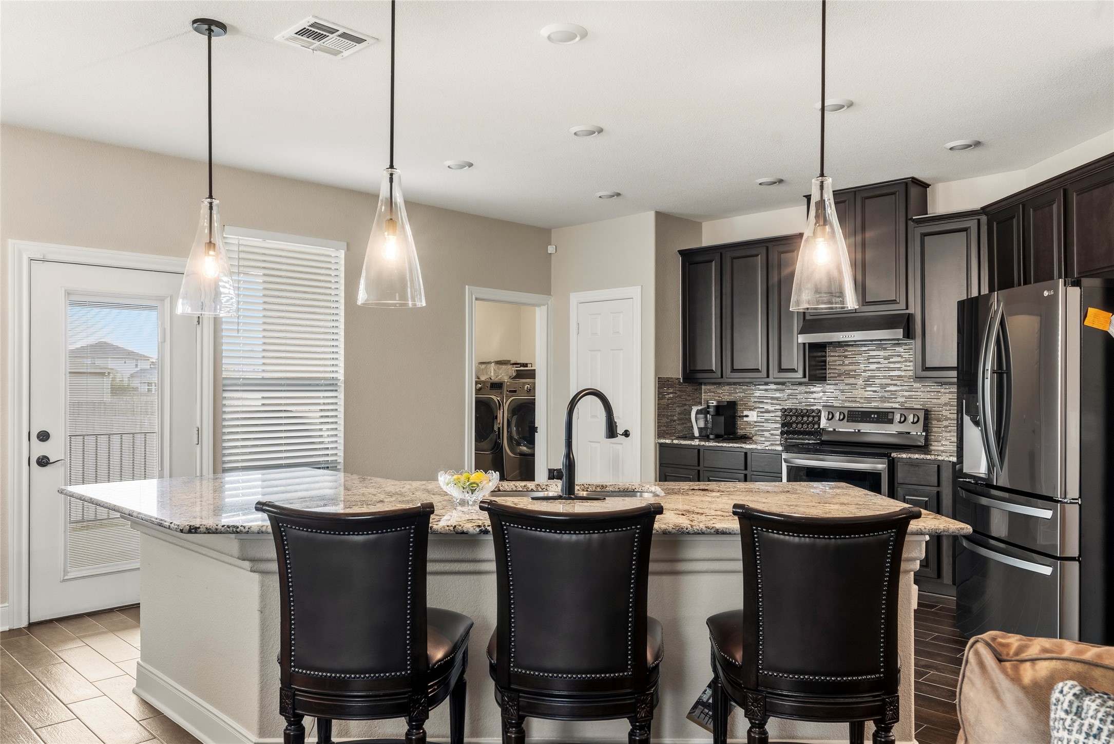 105 West Point Way Elgin, TX 78621 - Photo 19 of 27 Kitchen island featuring a granite countertop with seating, clear glass pendant lighting, dark wood cabinetry, a tiled backsplash, and stainless steel appliances