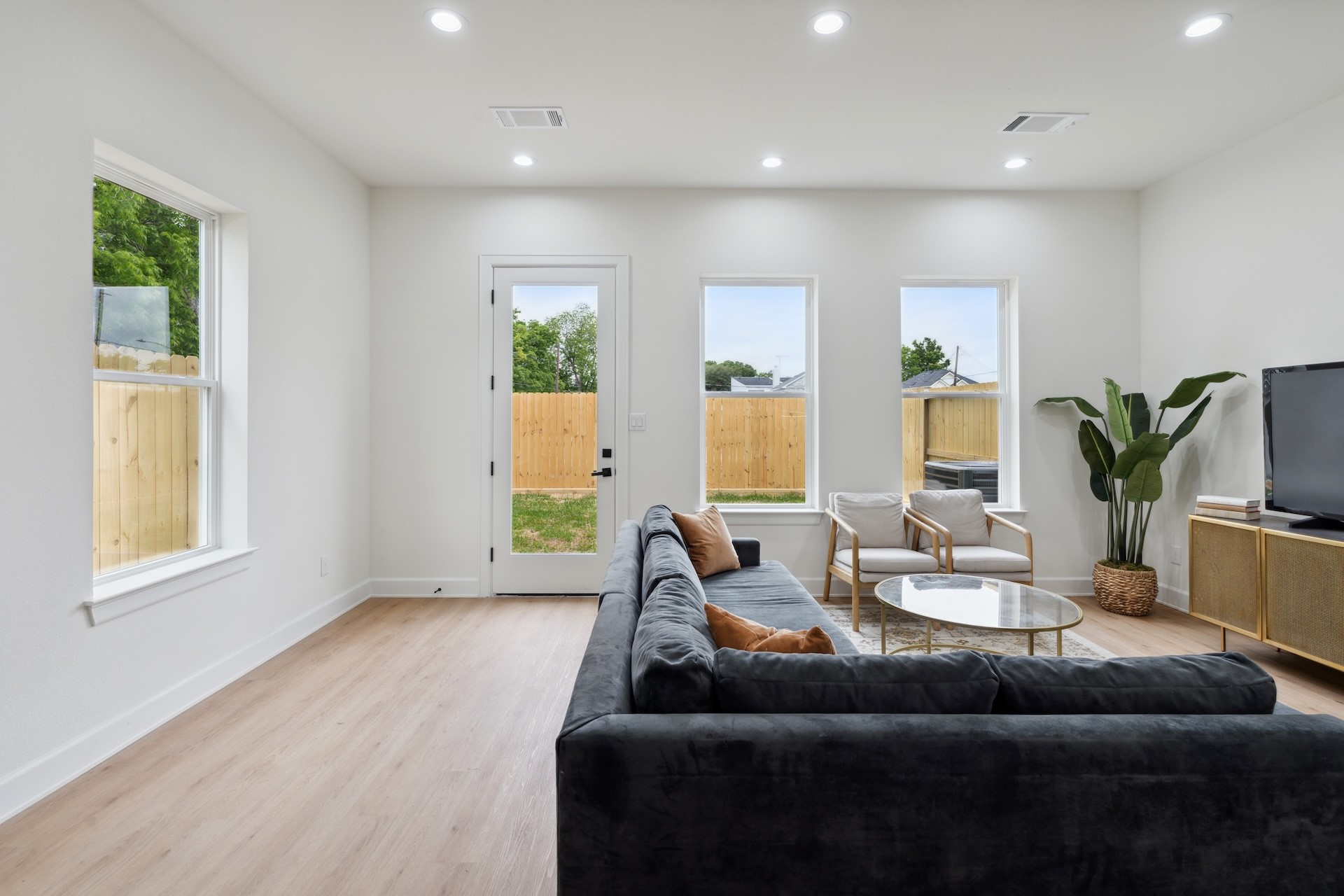 Open-concept living room — continuous wide-plank flooring, recessed LED lighting throughout, side-yard accent windows, and direct patio access.