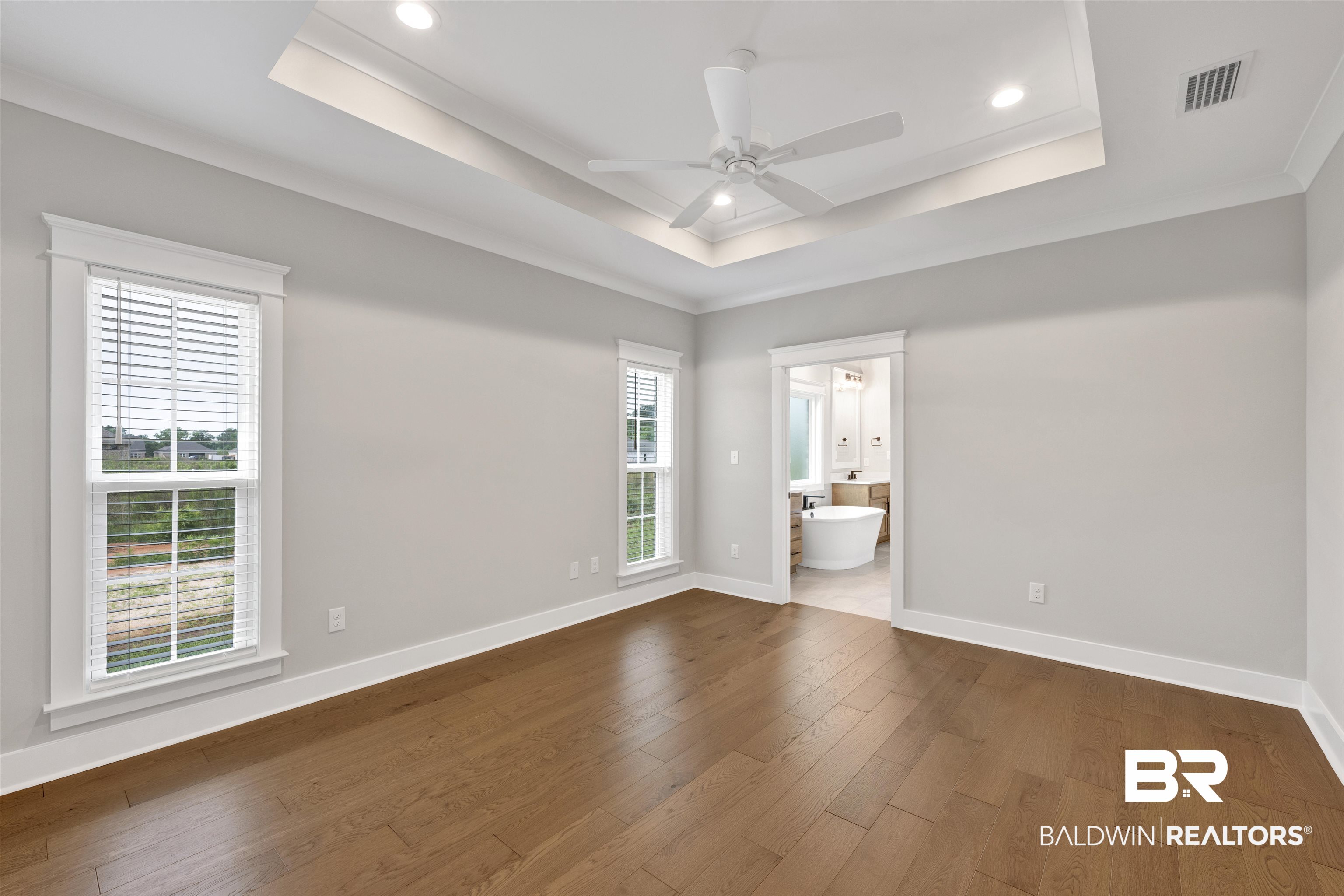 27371 Patch Pl Loop Daphne, AL 36526 - Photo 52 of 63 Bedroom featuring wood-finish flooring, two windows with white blinds, a tray ceiling with recessed lighting and a ceiling fan, and a doorway opening to a bathroom with a freestanding tub