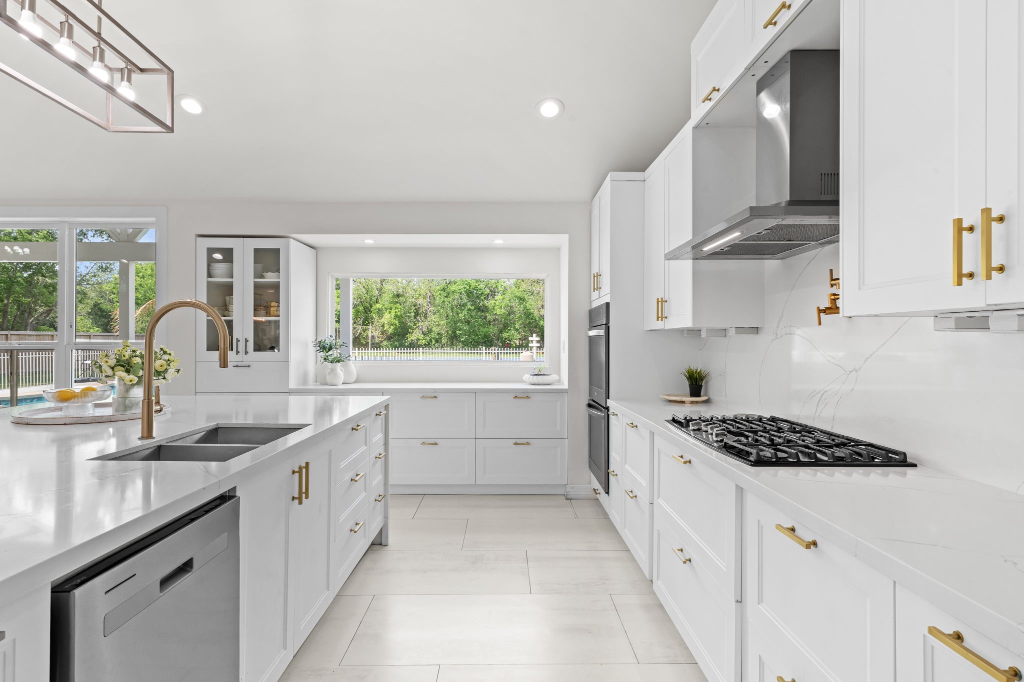 8800 Brae Acres Road Houston, TX 77074 - Photo 11 of 48 The range wall centers the kitchen's cooking zone — a gas range with a stainless steel hood above, full-height marble-look tile backsplash, and white shaker cabinetry with brushed gold hardware running the full perimeter on both sides. A large window above the sink brings the yard in from the far wall, keeping the space bright throughout the day.