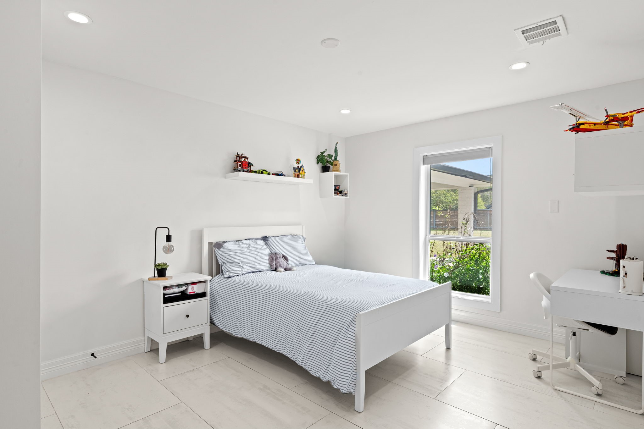 8800 Brae Acres Road Houston, TX 77074 - Photo 17 of 48 Large-format tile floors, recessed lighting, and a yard-facing window keep this secondary bedroom bright and livable. Floating shelves on the upper wall add display and storage space without taking up floor area — a small detail that earns its place in a room that works equally well as a guest room or home office.