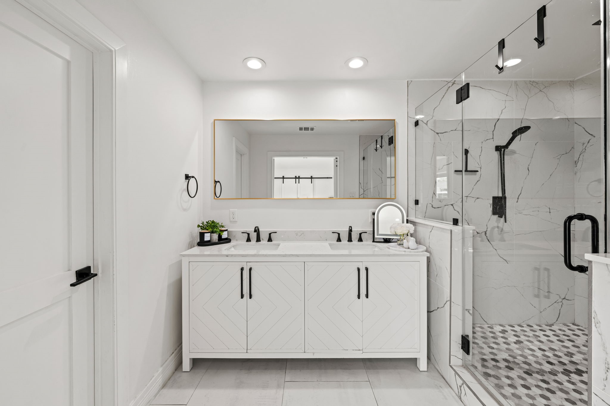 8800 Brae Acres Road Houston, TX 77074 - Photo 24 of 48 The primary bath delivers — a double vanity with herringbone-pattern cabinet fronts in a painted white finish and matte black hardware, topped with bright counters beneath a warm-toned rectangular mirror with a thin gold frame. Opposite, a frameless glass shower enclosure spans the full wall, tiled floor-to-ceiling in large-format porcelain with bold gray and white veining, finished with a matte black rain head and penny tile floor.