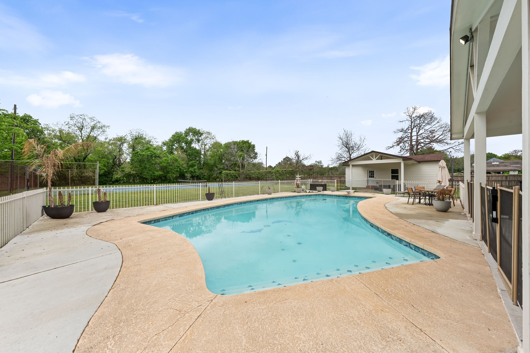 8800 Brae Acres Road Houston, TX 77074 - Photo 36 of 48 The freeform in-ground pool has a broad stamped concrete deck wrapping three sides and the covered patio running the full length of the fourth — the kind of setup that actually gets used. White perimeter fencing encloses the pool area without cutting off the sightlines to the open lawn and outbuildings beyond.
