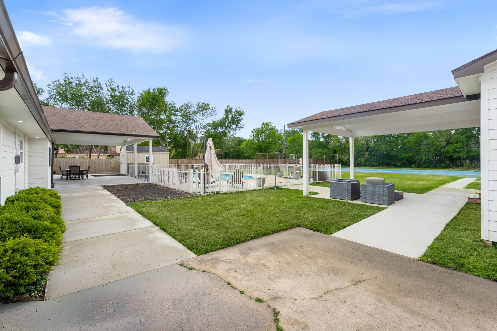 8800 Brae Acres Road Houston, TX 77074 - Photo 38 of 48 This is the view that puts the full outdoor program in one frame — covered patio on the left, guest quarters on the right, pool in the center background, and the tennis court visible at the far right edge. Concrete walkways connect it all across a manicured lawn, with the mature tree line along the rear fence providing natural enclosure without closing anything off.