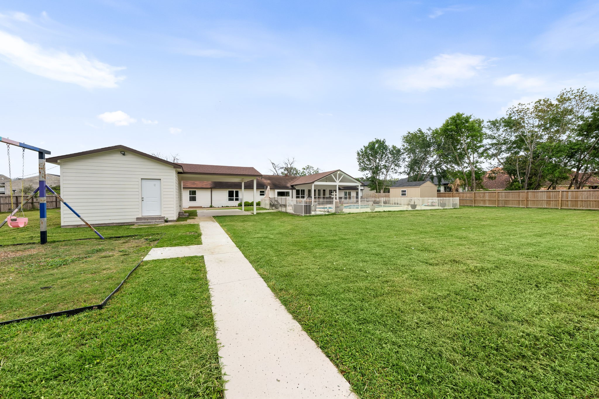 8800 Brae Acres Road Houston, TX 77074 - Photo 41 of 48 A concrete walkway runs the length of the rear yard alongside the storage building, connecting the far end of the lot to the pool enclosure and covered patio beyond. The open lawn on both sides and the main house visible at the far end give a clear sense of how much usable ground the property holds between the two.