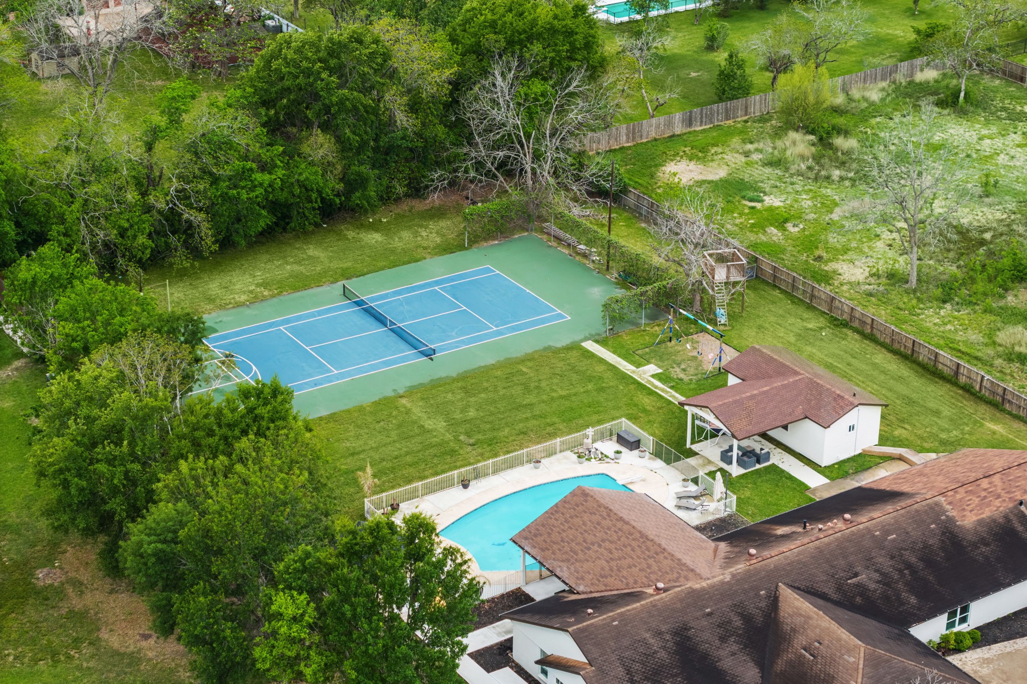 8800 Brae Acres Road Houston, TX 77074 - Photo 44 of 48 An aerial that captures the two marquee outdoor amenities in a single frame — the freeform pool with its white perimeter fence in the lower portion, and the full tennis court beyond it at the rear of the lot. Mature tree coverage wraps the property on multiple sides, giving the entire property a naturally enclosed quality that the ground-level photos can only partially communicate.