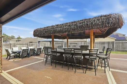 24714 Longwood Forest Drive Spring, TX 77373 - Photo 17 of 21 This photo showcases a spacious outdoor patio with a large tiki-style bar and ample seating, perfect for entertaining. The area is well-paved and surrounded by a privacy fence, offering a relaxed and inviting atmosphere.