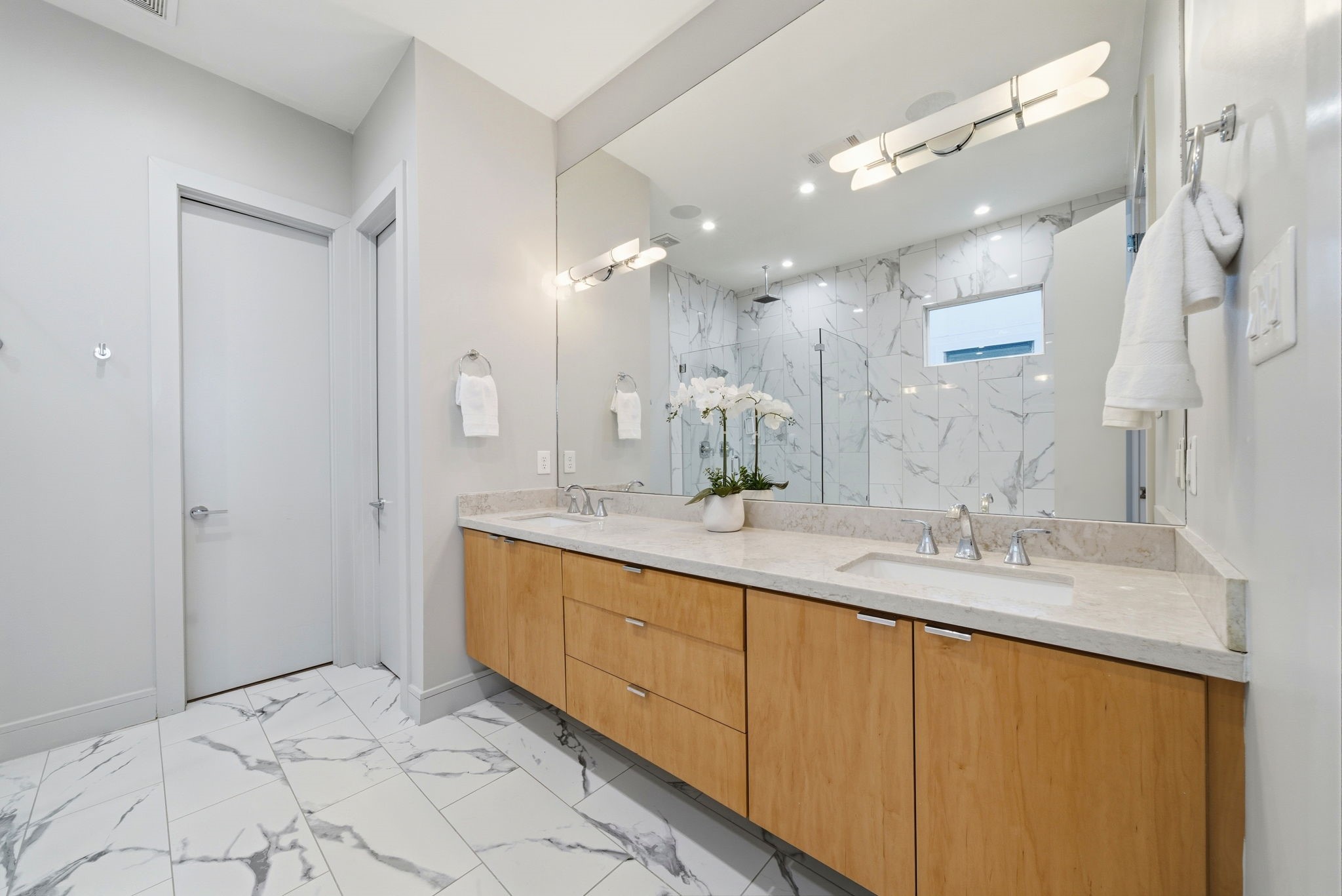 1405 Vermont Street, Unit A Houston, TX 77006 - Photo 31 of 46 Dual sink vanity with warm wood cabinetry and quartz countertops anchors this renovated primary ensuite bath. Large mirrors reflect the designer finishes and a spacious glass-enclosed shower featuring floor-to-ceiling marble-style tile. Marble-inspired porcelain tile floors flow seamlessly through the space, where a transom window and recessed LED lighting ensure a bright, luxury atmosphere. This spa-inspired retreat offers both a walk-in shower and easy access to the expansive walk-in closet.