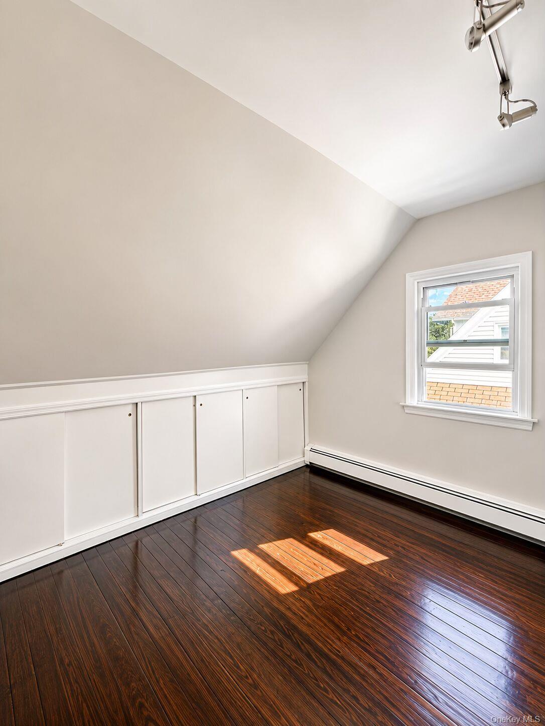 79 Division Street, Unit 3 New Rochelle, NY 10805 - Photo 8 of 17 Second bedroom featuring wood-finish flooring, built-in lower cabinetry with paneling, a single window with white trim, a sloped ceiling, and baseboard heating.