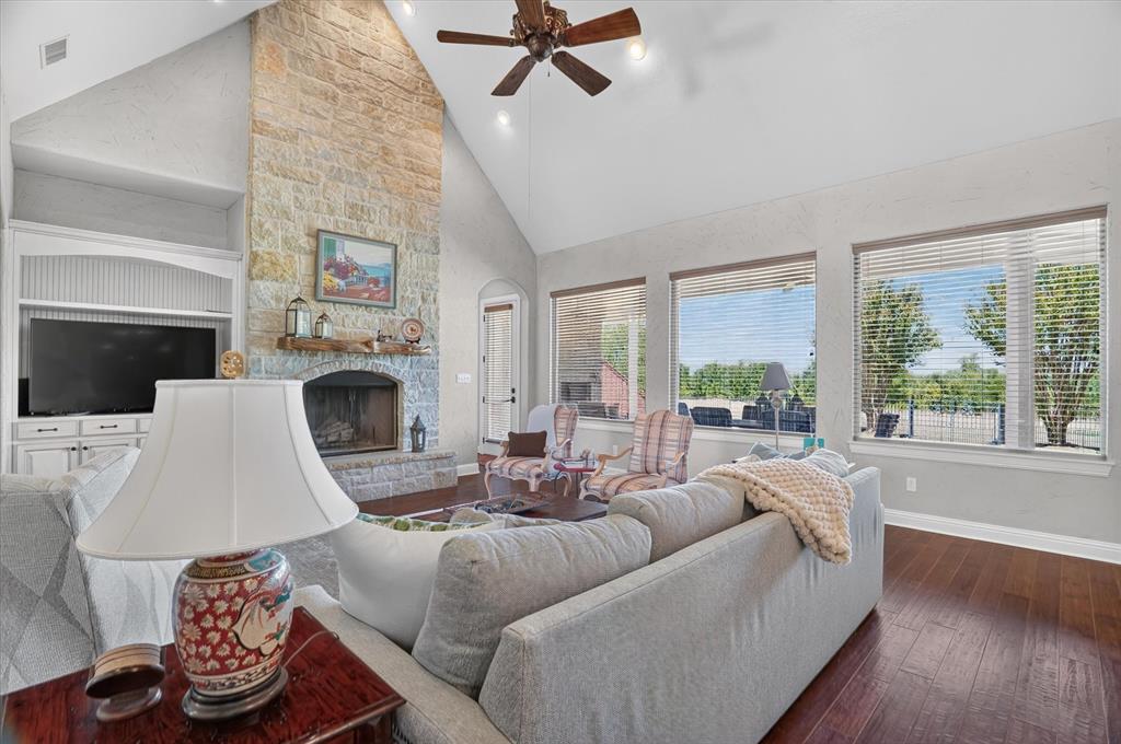 Bright, vaulted great room featuring a wood-burning fireplace with stone surround, hardwood flooring, and abundant natural light from multiple large windows and glass doors