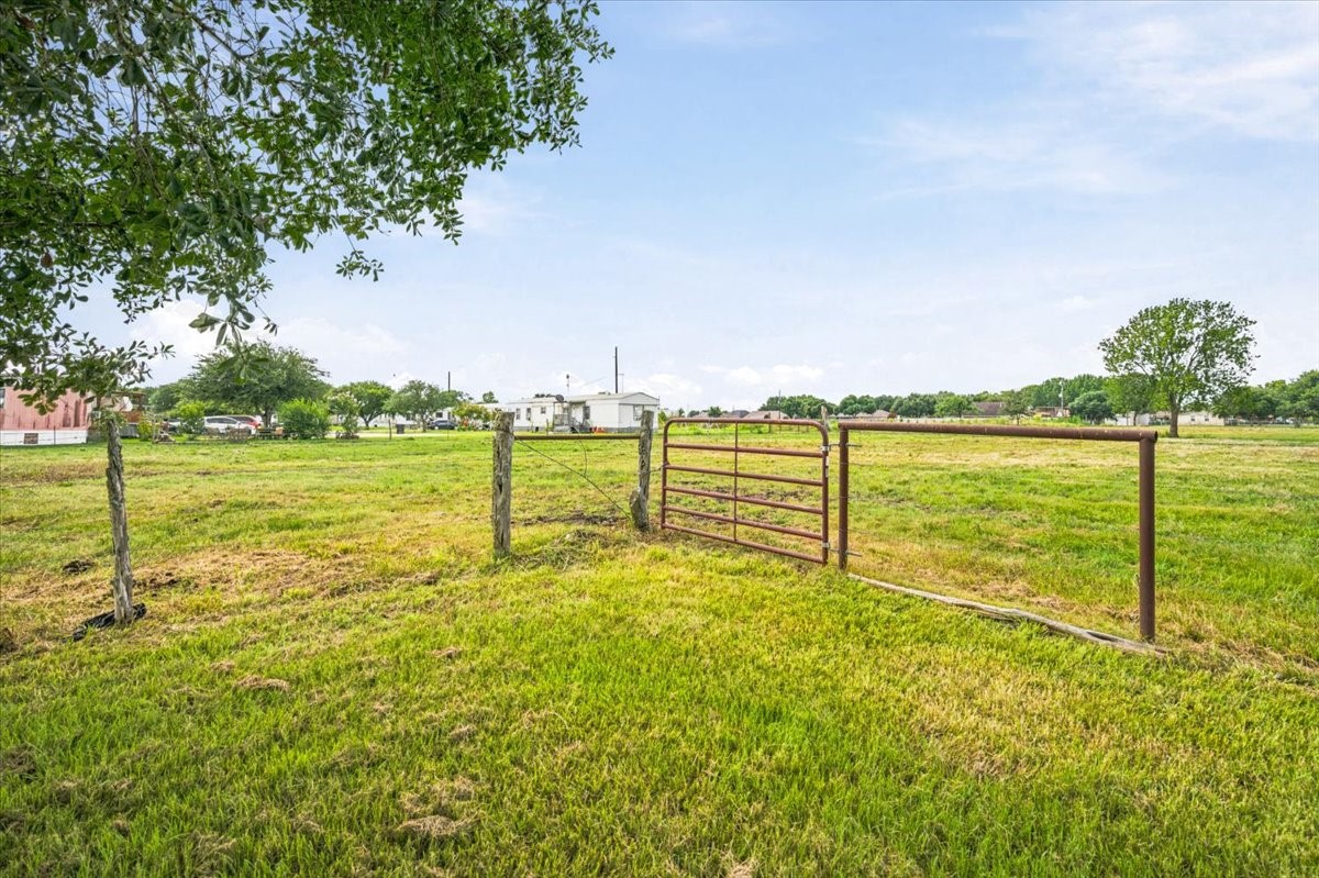 765 Norcross Road Wallis, TX 77485 - Photo 28 of 42 This photo showcases a spacious, open grassy field with a metal gate and rustic wooden posts. It's bordered by trees and offers a view of a few distant houses, perfect for someone seeking a rural or semi-rural setting.