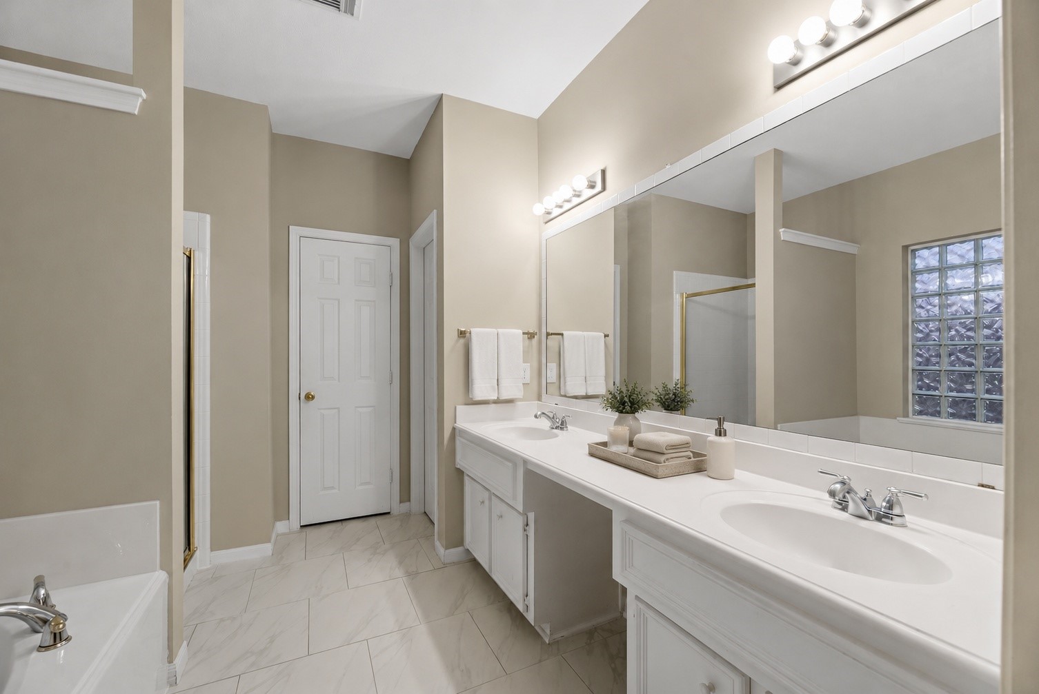 5724 Shady Hollow Court Rosharon, TX 77583 - Photo 12 of 27 Spacious primary suite bathroom featuring tall ceilings, dual vanities, expansive mirror space, and bright vanity lighting. The layout offers excellent functionality with separate sink areas, generous countertop space, a soaking tub, separate shower, and a glass-block window that brings in natural light while maintaining privacy. Light tile flooring and neutral tones create a clean, airy feel with great potential for a calm, spa-inspired retreat.