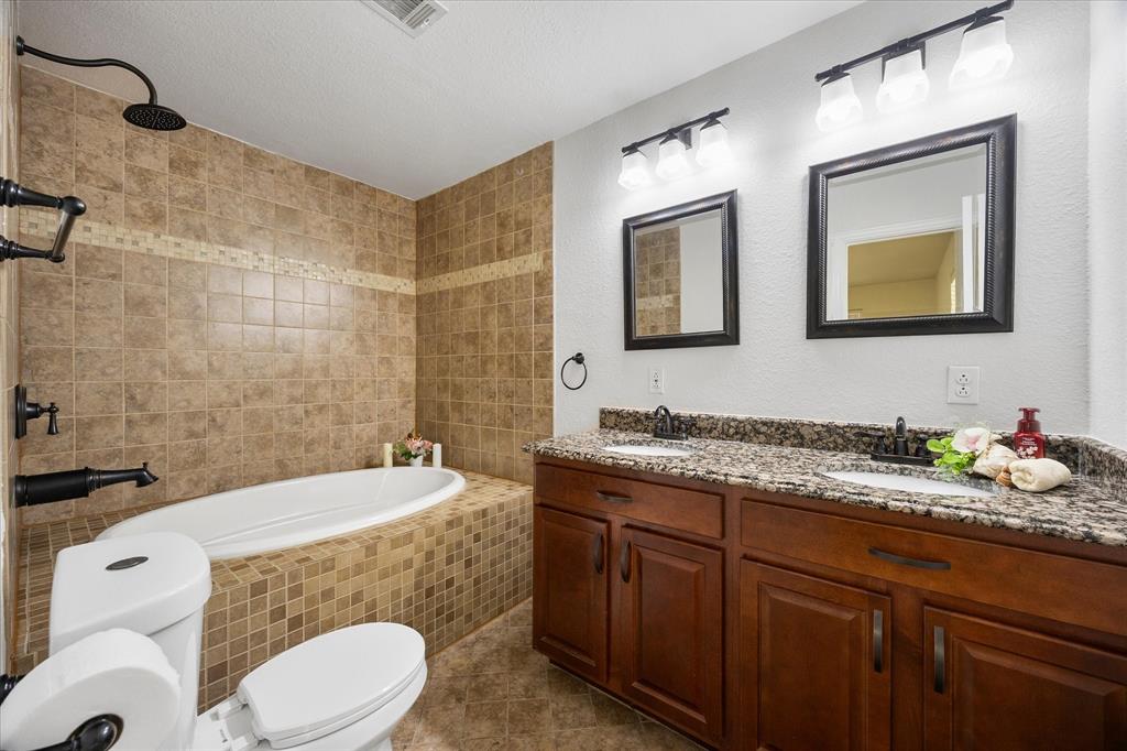 925 Lake North Road Rowlett, TX 75089 - Photo 19 of 37 Soaking tub with tiled surround, dual vanity with undermount sinks, stone countertop, wood cabinetry, and wall-mounted mirrors with lighting