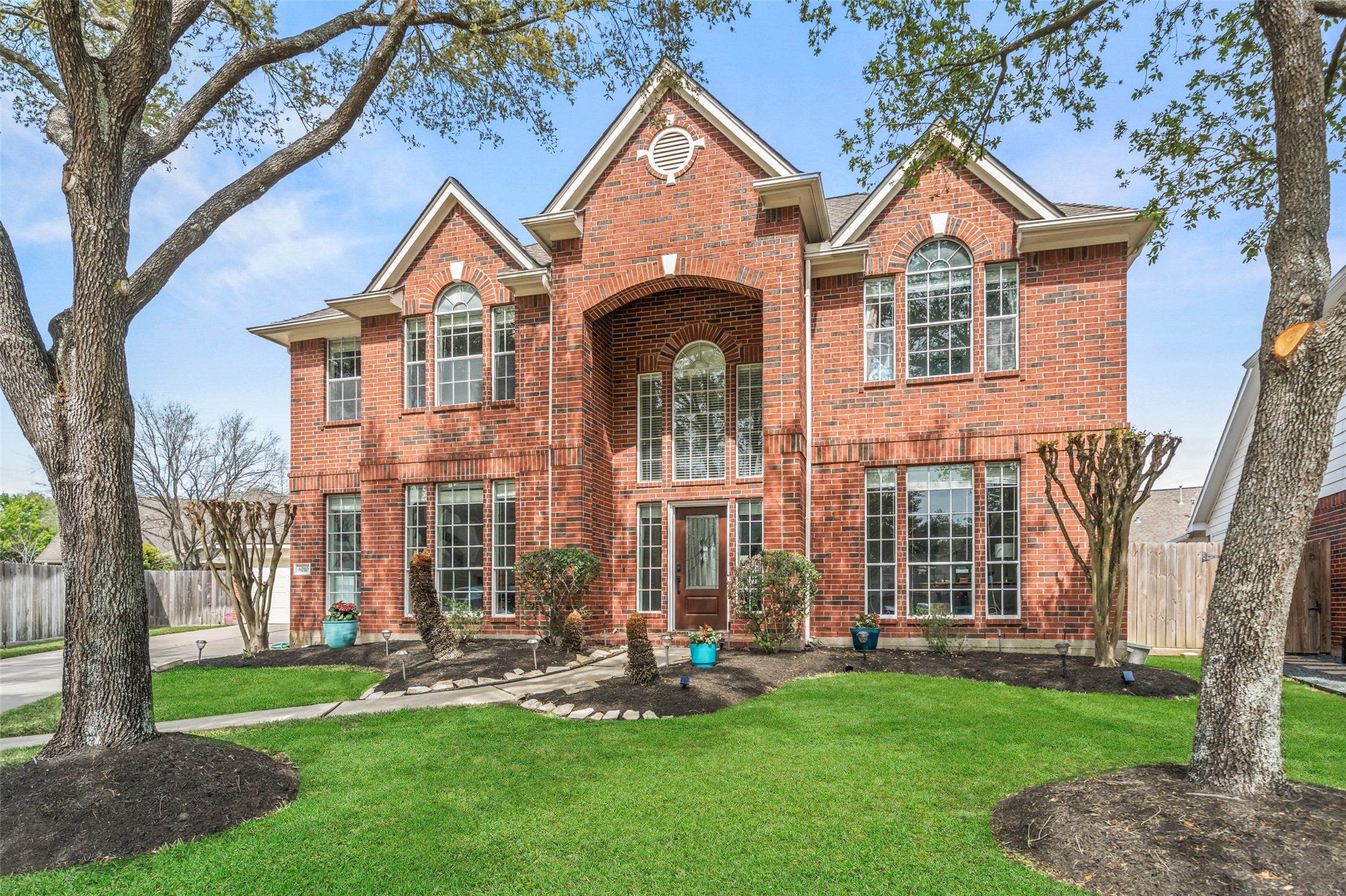 This elegant two-story brick home features a striking facade with arched windows, a welcoming entrance, well-maintained landscaping, and mature trees, offering both curb appeal and a cozy atmosphere.