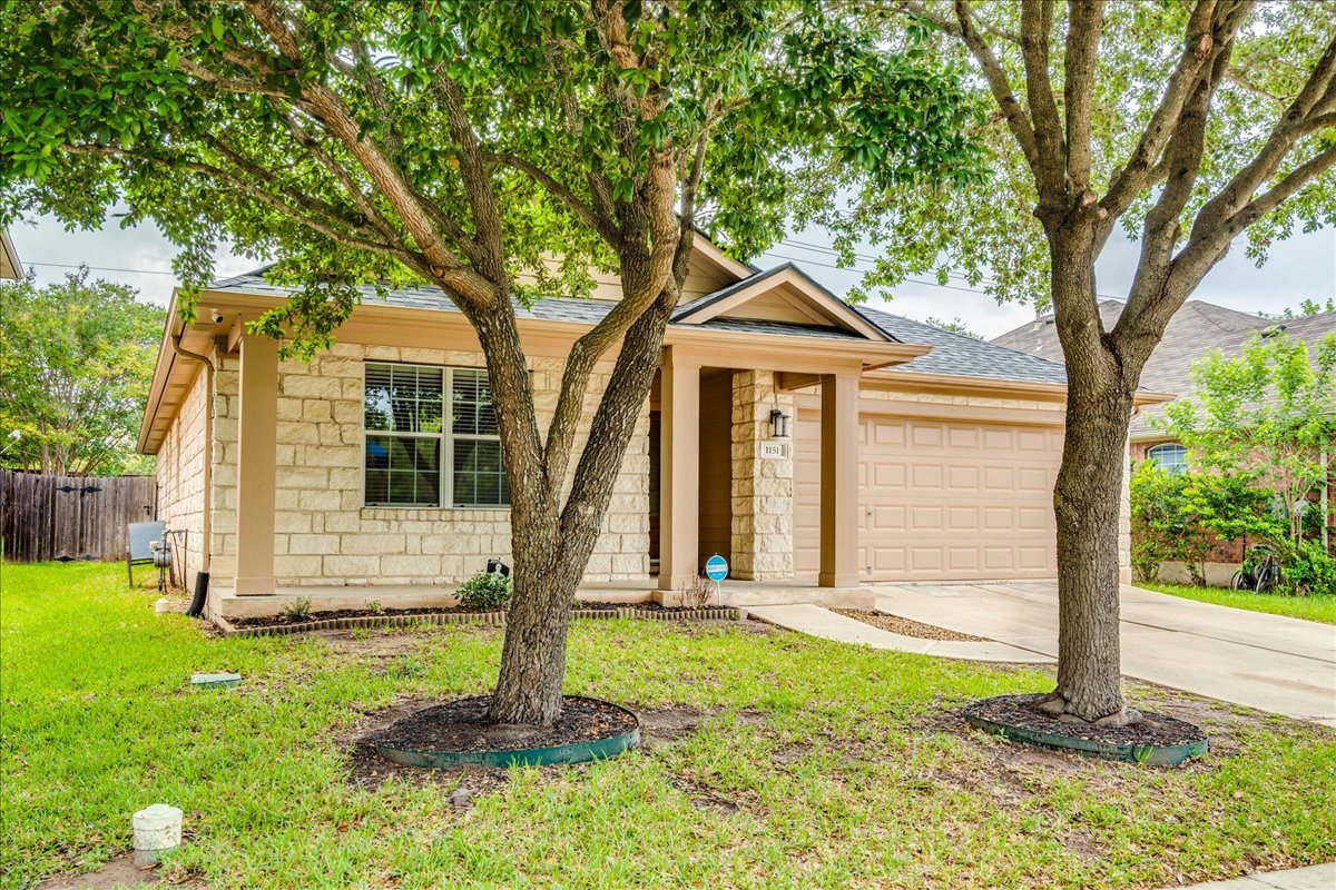 1151 Stone Rim Loop Buda, TX 78610 - Photo 34 of 35 The property features a stone facade, an attached garage, a covered entryway, and a well-maintained lawn with mature trees
