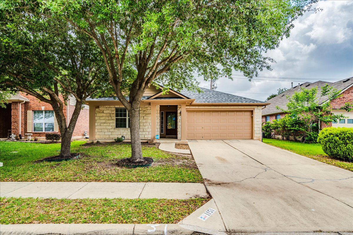 1151 Stone Rim Loop Buda, TX 78610 - Photo 35 of 35 The property features a stone facade, an attached garage, a paved driveway, and a well-maintained lawn with mature trees