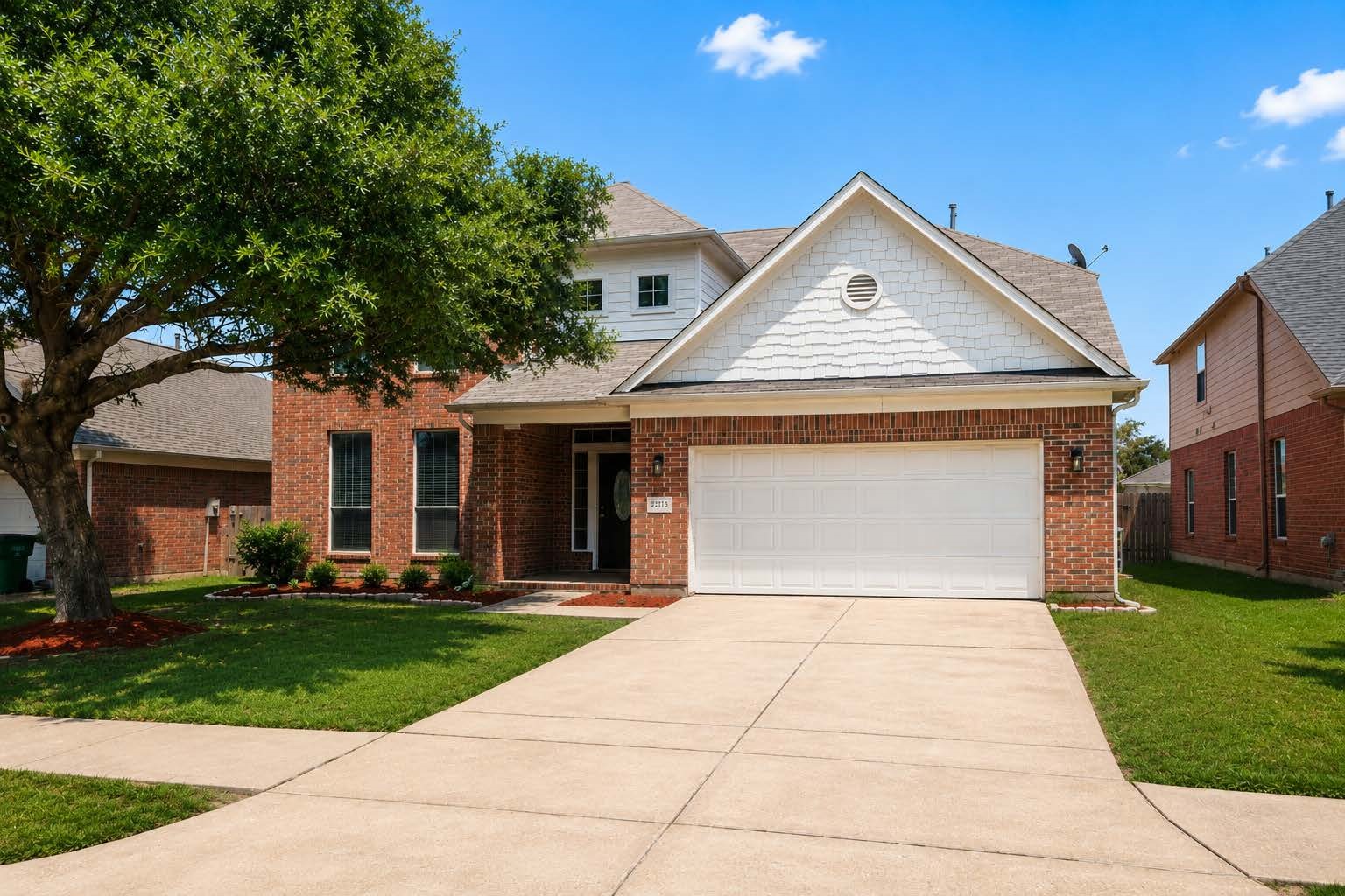This photo shows a charming two-story brick house with a two-car garage. The front features a mature tree and double wide driveway.
