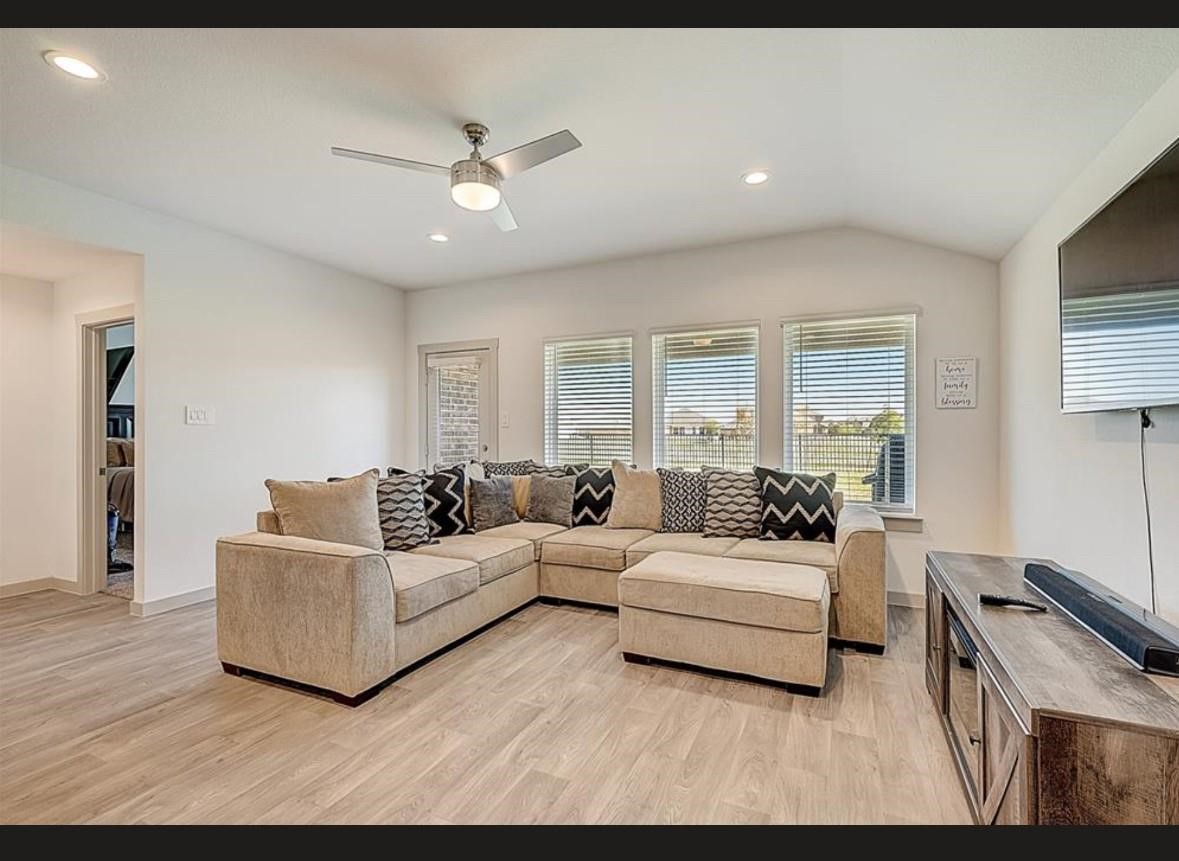 1246 Truckee River Drive Rosharon, TX 77583 - Photo 11 of 23 Bright and modern living room featuring a large sectional sofa, sleek ceiling fan, and mounted TV. Ample natural light streams through multiple windows, highlighting the light wood flooring.