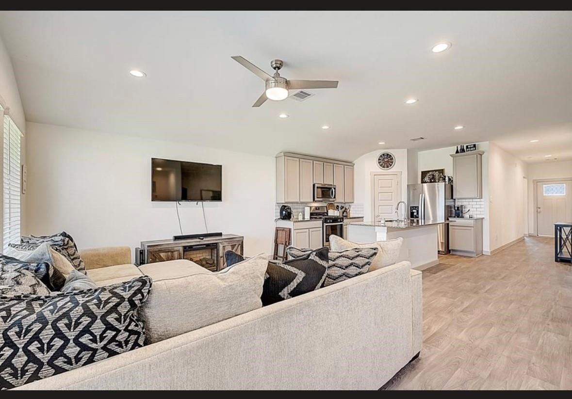 1246 Truckee River Drive Rosharon, TX 77583 - Photo 12 of 23 This photo showcases an open-concept living area with a modern kitchen and cozy living room. The space features light wood flooring, a ceiling fan, and recessed lighting. The kitchen includes stainless steel appliances, an island, and ample cabinetry, while the living room has a large sectional sofa and wall-mounted TV. Perfect for entertaining and family gatherings.