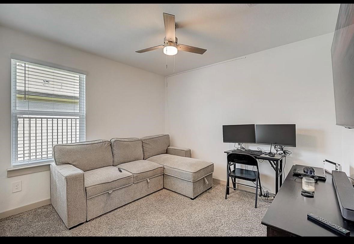 1246 Truckee River Drive Rosharon, TX 77583 - Photo 13 of 23 Cozy room with neutral tones featuring a comfortable L-shaped sofa, dual-monitor desk setup, and ceiling fan. Carpeted flooring adds warmth, while a large window provides natural light. Ideal for a home office or relaxation space.