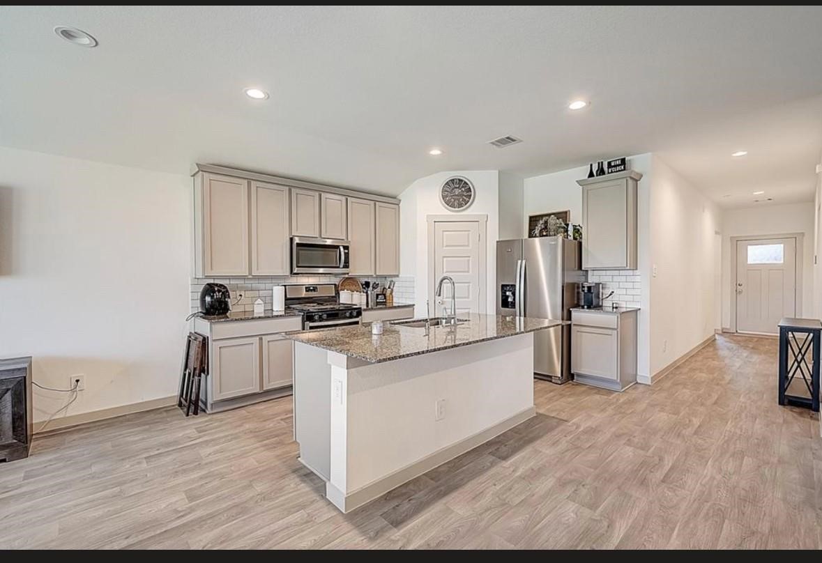 1246 Truckee River Drive Rosharon, TX 77583 - Photo 4 of 23 This photo shows a modern kitchen with sleek gray cabinets, stainless steel appliances, and a central island with a granite countertop. The space is open and bright, featuring wood-look flooring and recessed lighting. A hallway leads to a door, adding to the open layout.