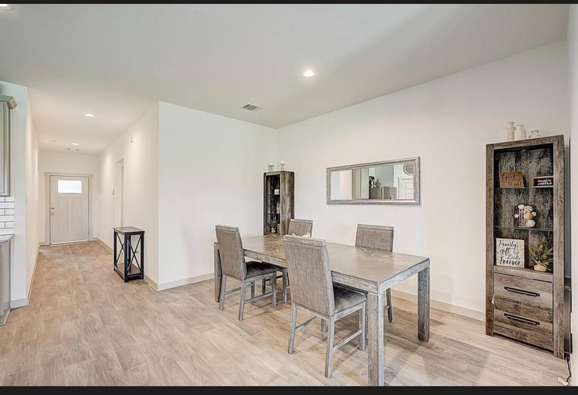 1246 Truckee River Drive Rosharon, TX 77583 - Photo 9 of 23 This photo showcases a modern dining area with a wooden table and chairs, complemented by light wood flooring. The space is accented with a large wall mirror and two rustic shelves, creating a cozy yet contemporary atmosphere. The room is well-lit and leads to a welcoming entryway.