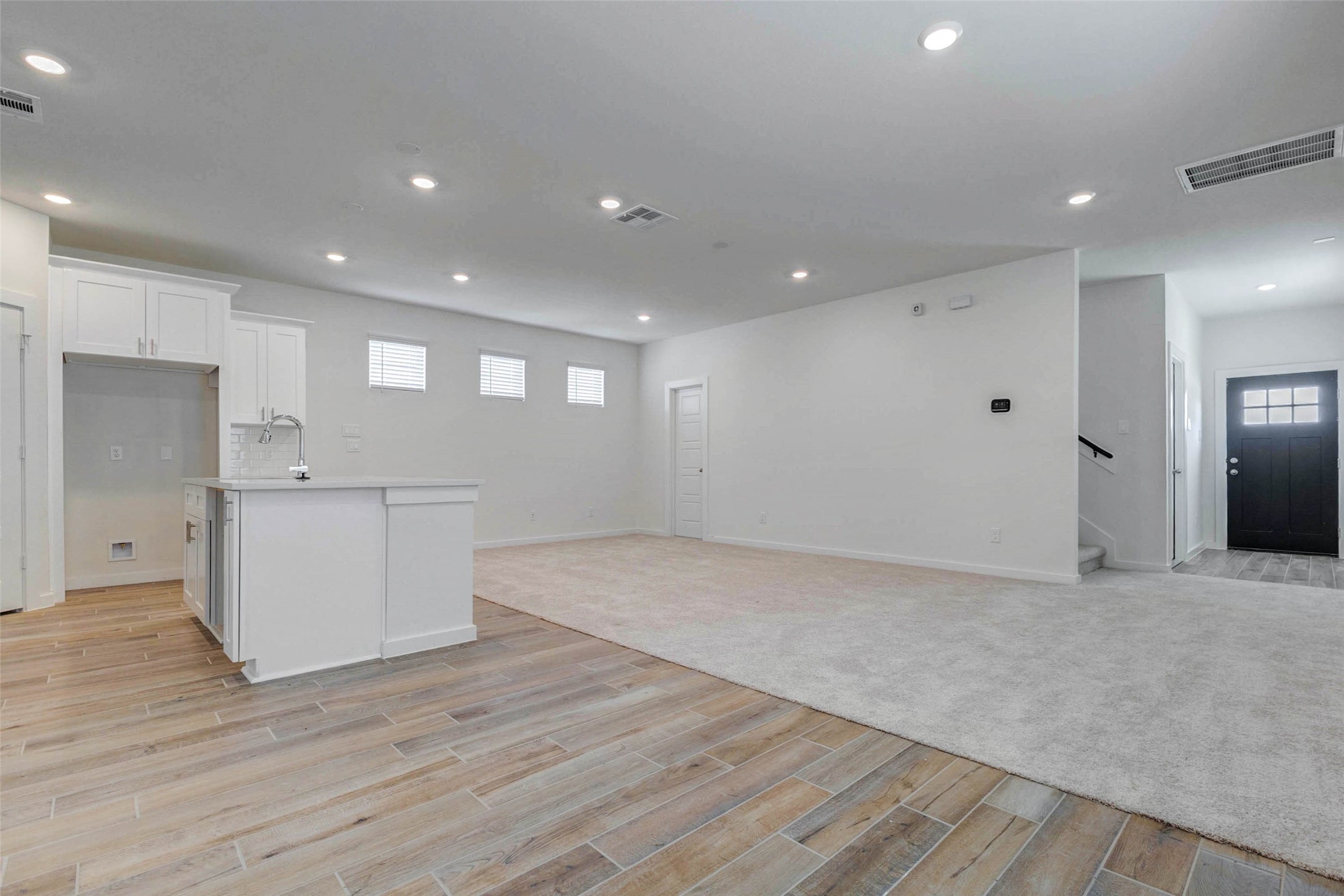 6471 Escondido Drive Rosharon, TX 77583 - Photo 12 of 33 Wide open view from the kitchen into the living area, showing the island, white cabinetry, wood-look tile, carpeted living space, recessed lighting, and front entry beyond. The layout provides easy flow for entertaining while keeping the main living spaces connected.