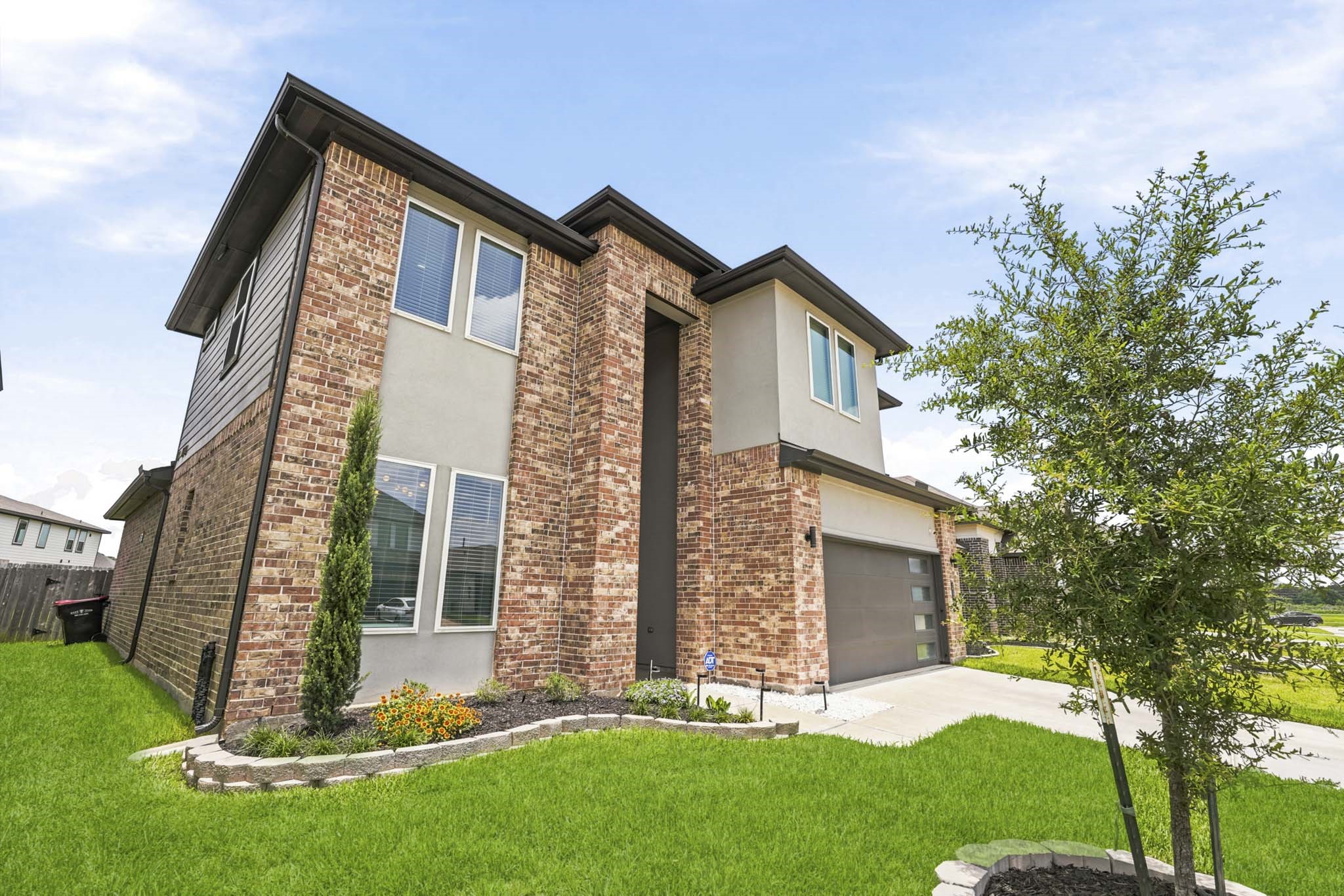 22522 Modica Rift Lane Katy, TX 77449 - Photo 6 of 38 Every angle of this home delivers. The corner view reveals the full architectural depth of the design — rich brick wrapping the exterior, tall vertical windows flooding the interior with light, and carefully curated landscaping with flowering plants and Italian cypress that add warmth and character to the curb appeal.
